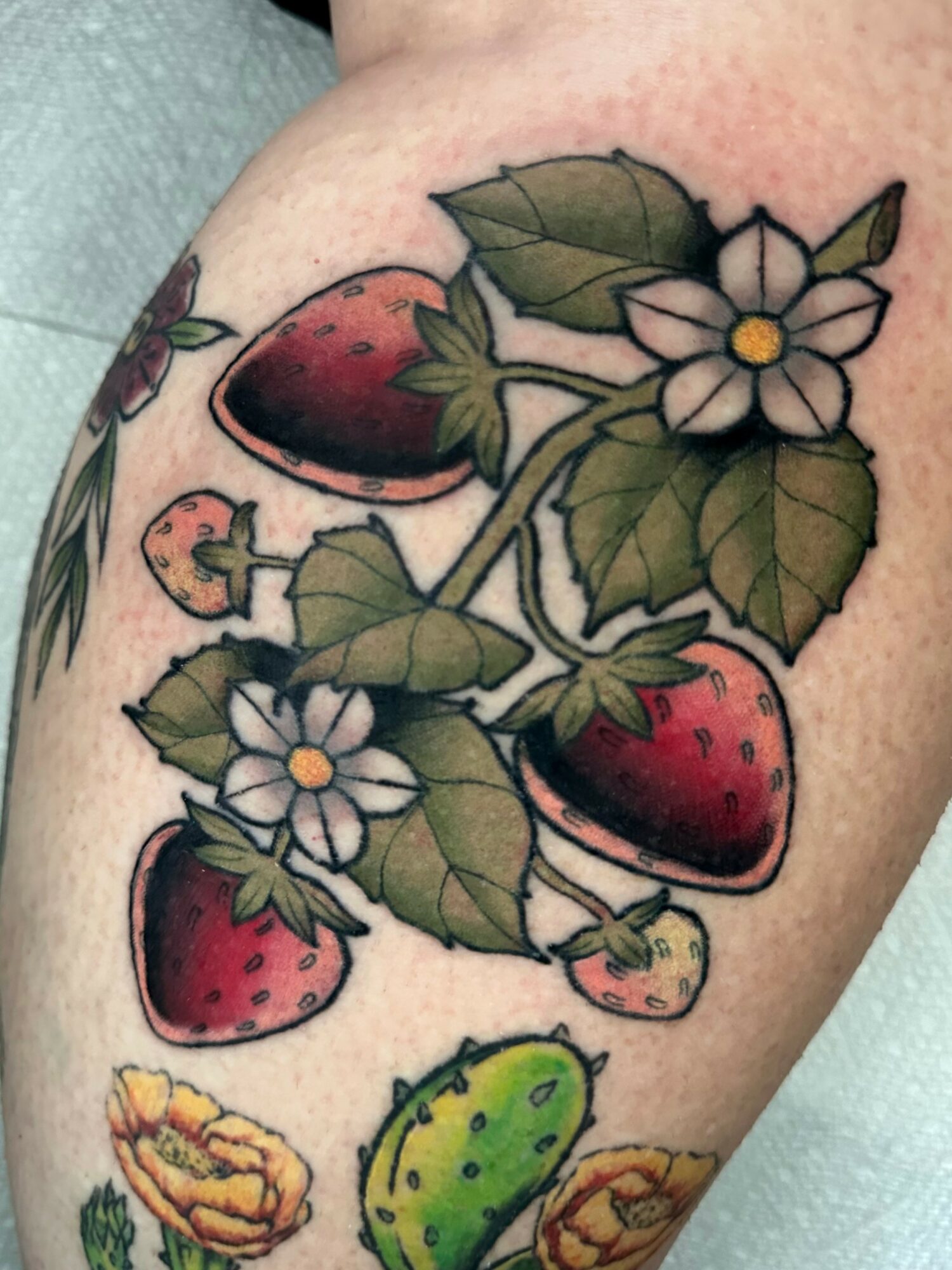 Colorful strawberry plant tattoo with strawberries, leaves, and flowers on skin.