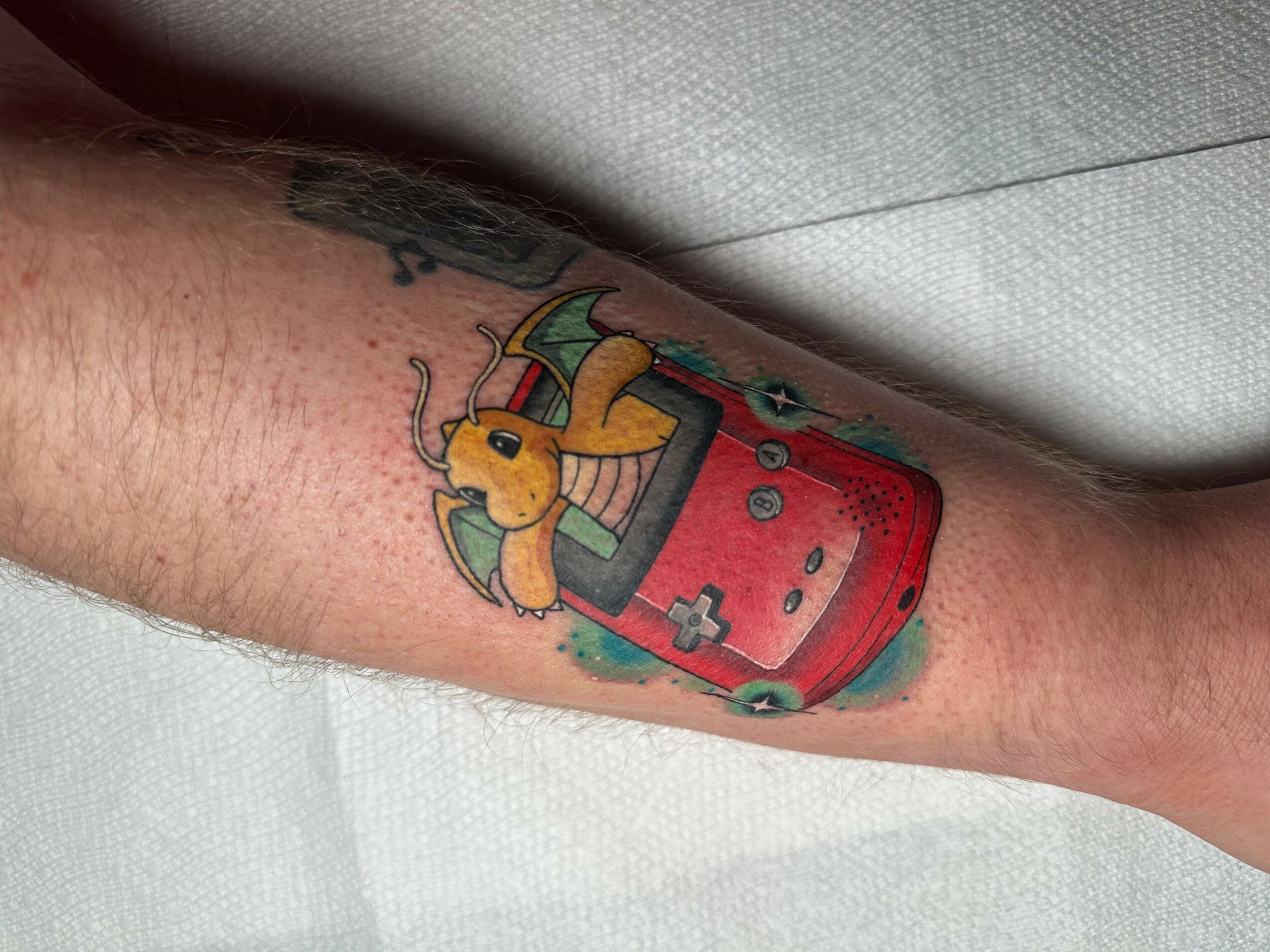 Tattoo of a yellow cartoon creature with wings and a tail, inside a red handheld gaming device, on person's arm