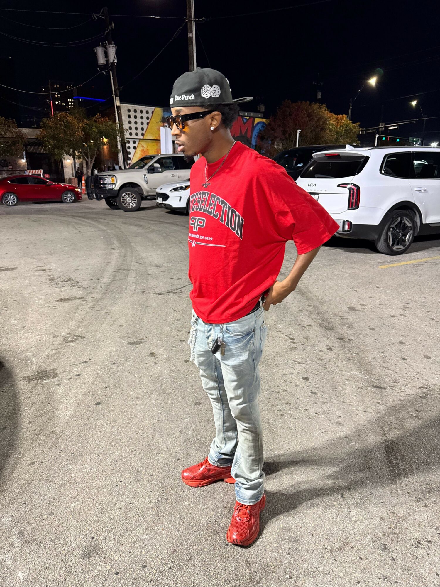 Person in red shirt and light jeans walking on a parking lot at night, with cars in the background.