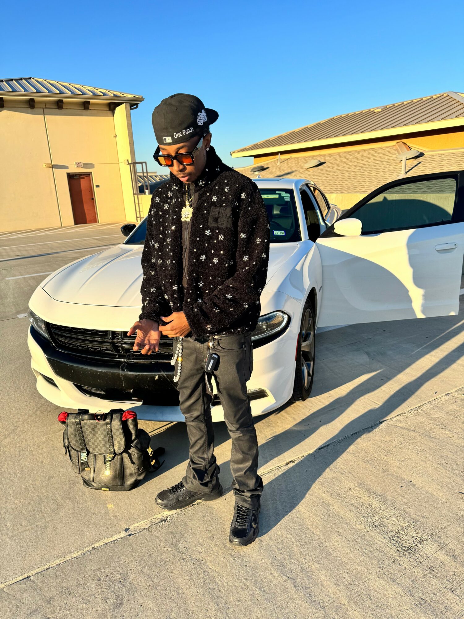Young man standing next to a white sports car outdoors, wearing a black jacket, sunglasses, and a cap, with a backpack on the ground.