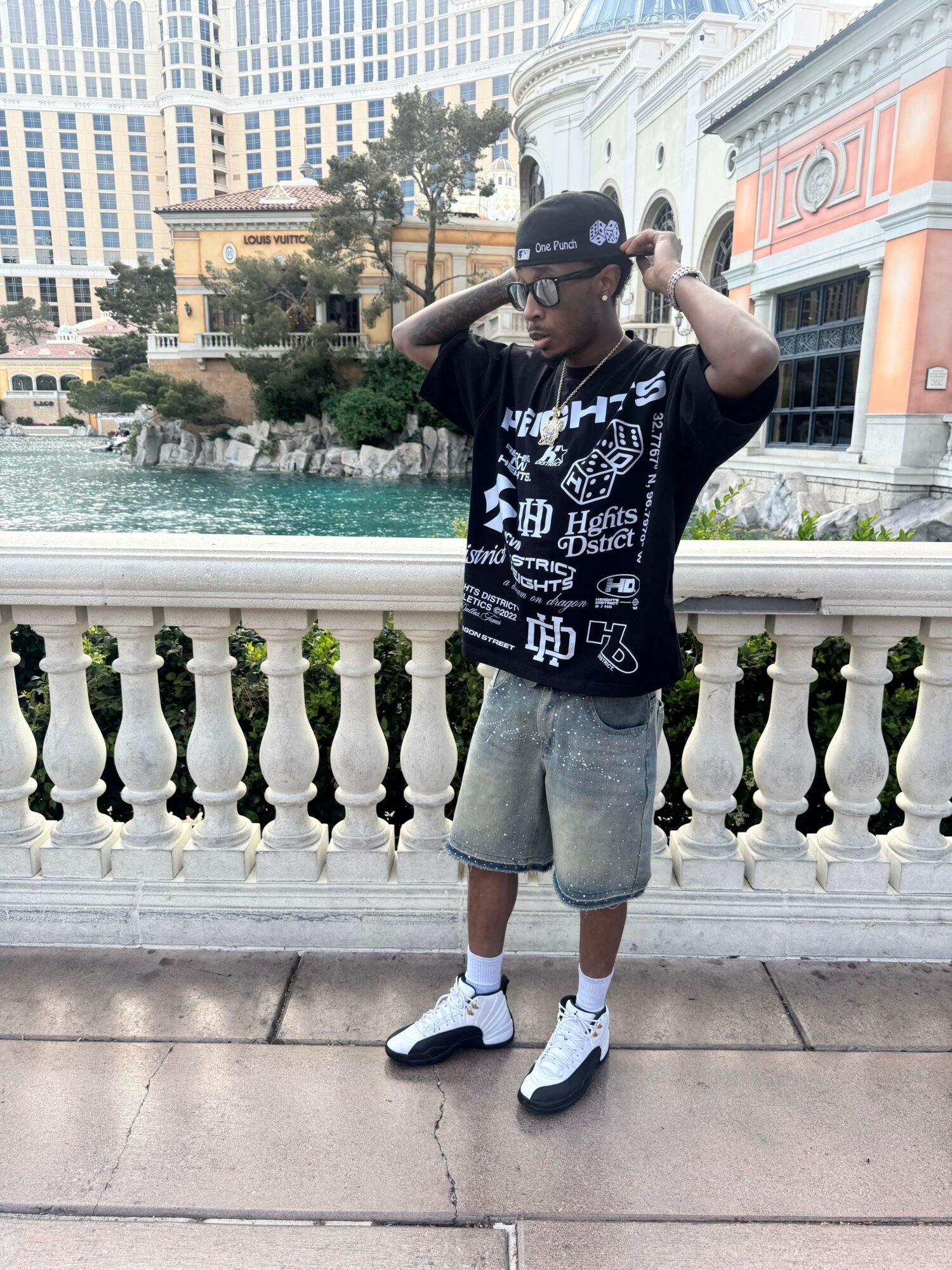 Person standing outdoors on a sidewalk, wearing a black T-shirt, gray shorts, white sneakers, and a cap, holding a phone to their ear.