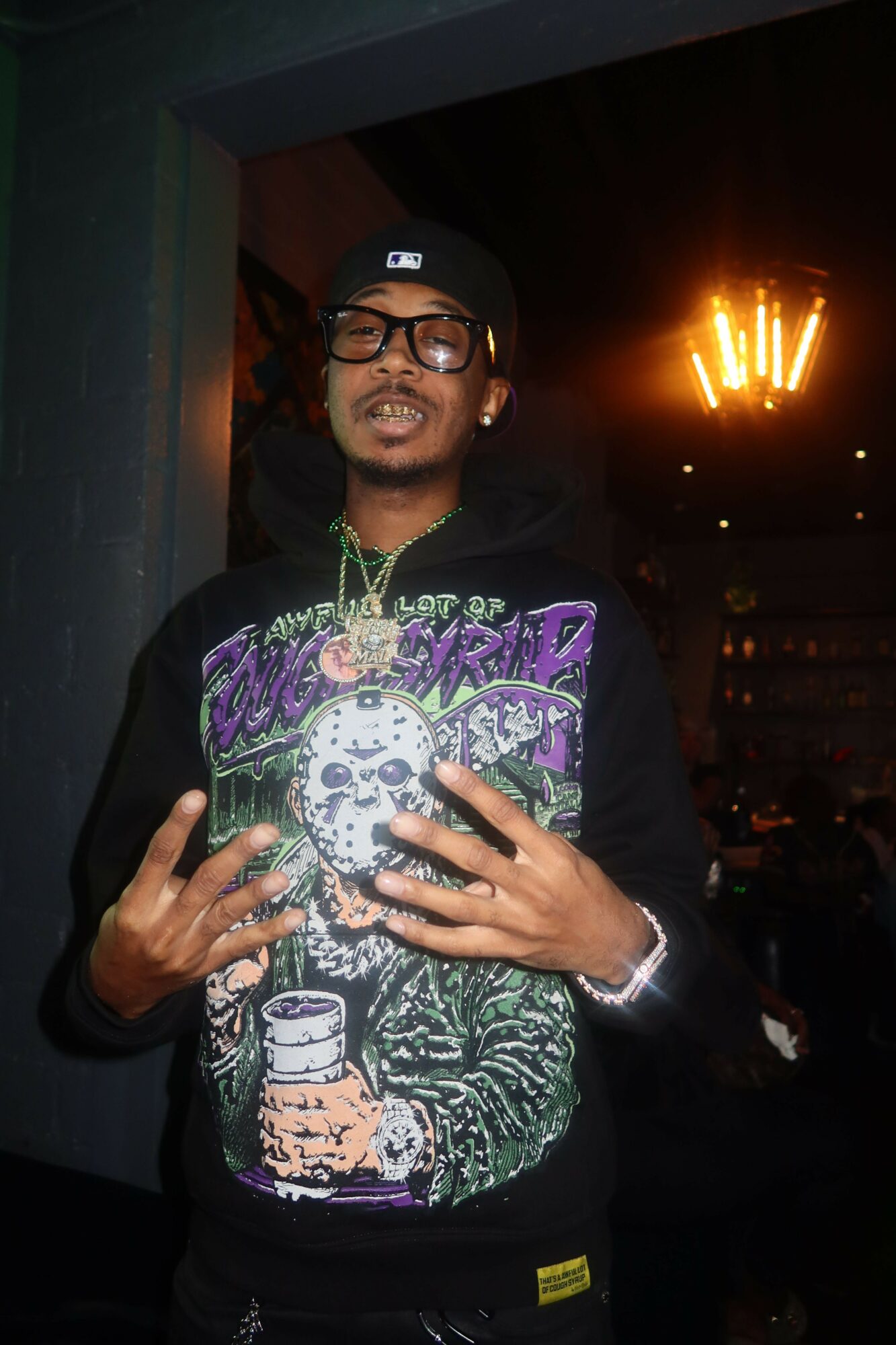 Person wearing glasses and a black hoodie with colorful graphic, holding a phone, in a dark urban setting at night.