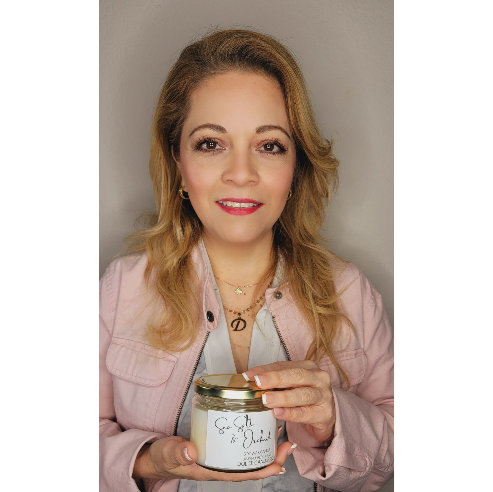 Woman with wavy blonde hair holding a jar with a label, smiling, against a plain background.