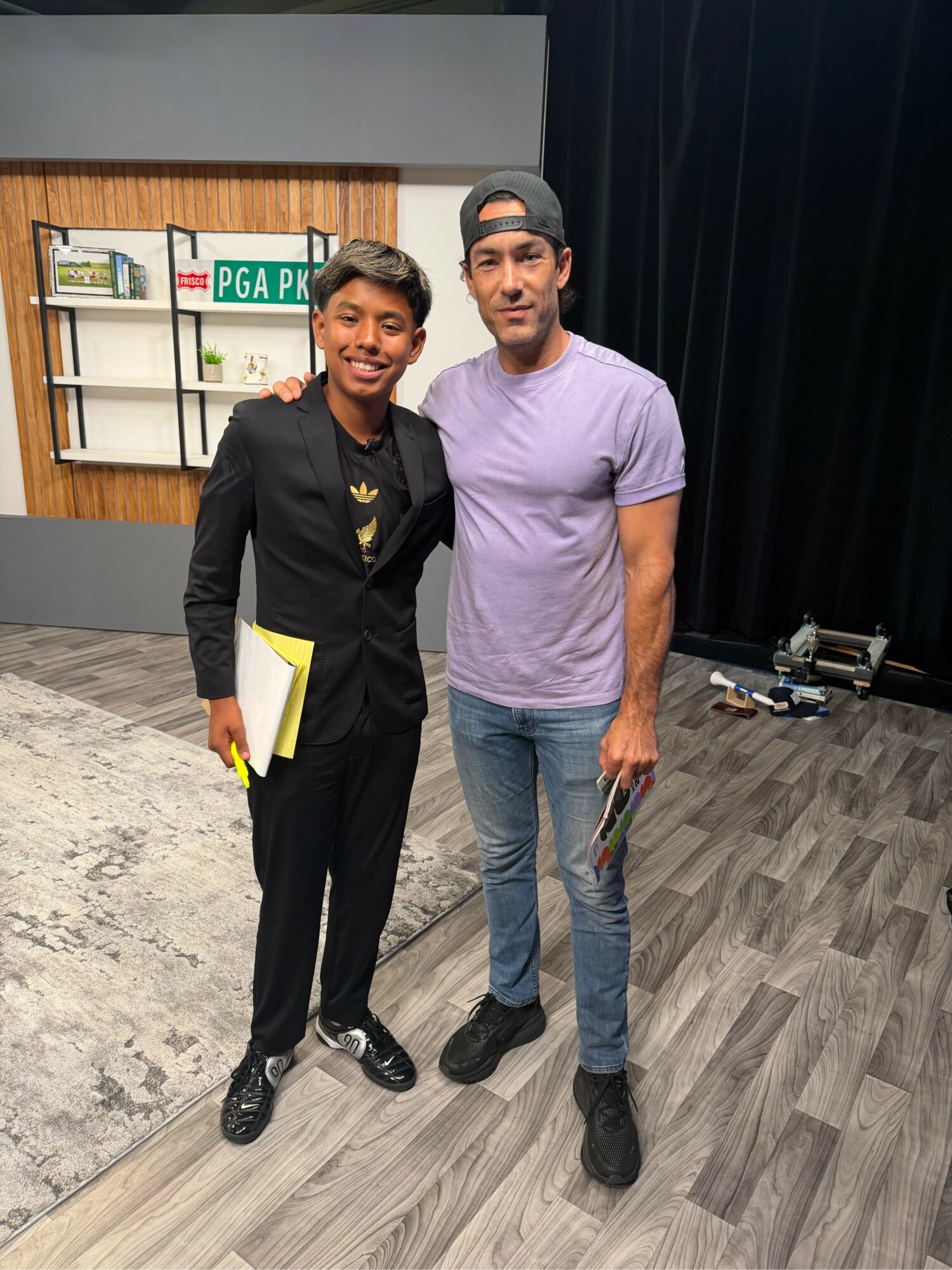 Two men standing together indoors, one in a black suit holding papers, the other in a purple t-shirt and jeans, smiling.