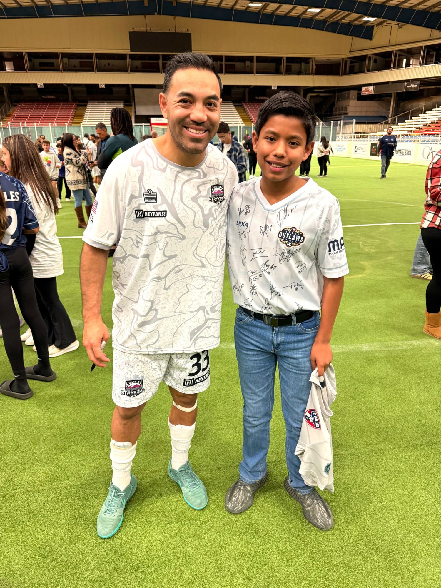 Two people standing on a sports field, smiling, with a stadium background and other people around.