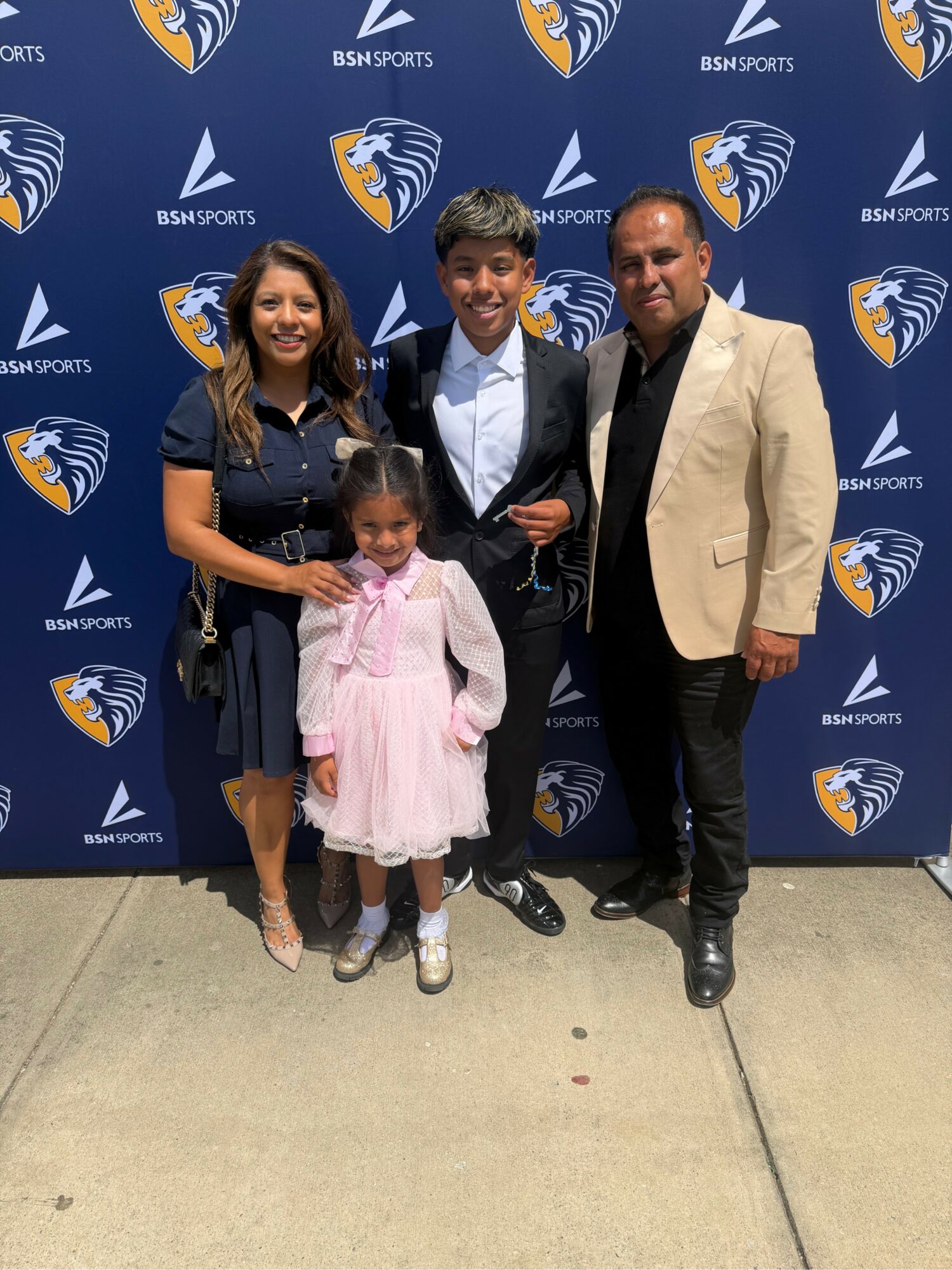 Four people, two children and two adults, stand in front of a blue backdrop with sports logos, smiling for a photo.