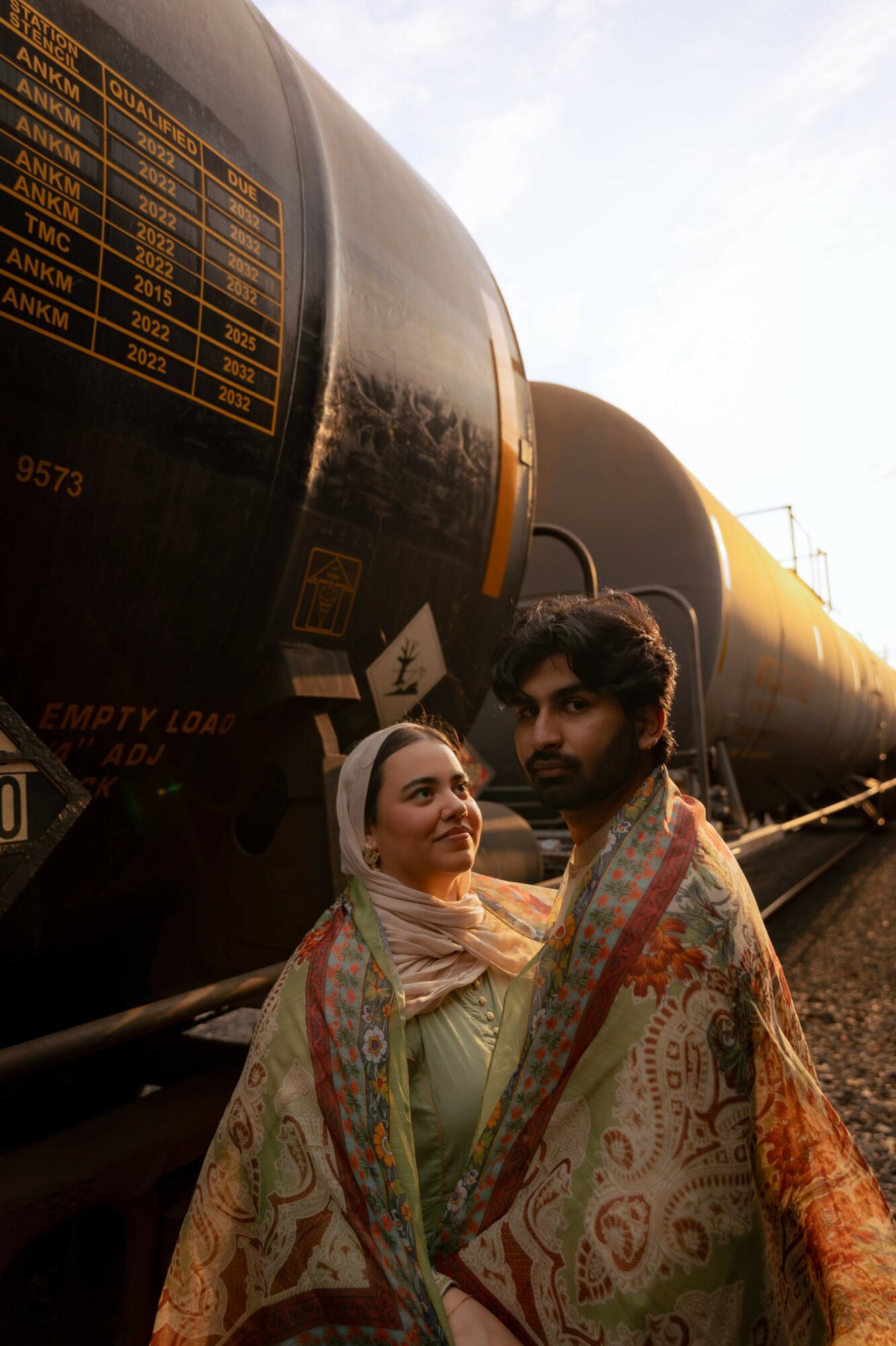Two people standing near black cylindrical tanks, one woman wearing a headscarf and the other man with a shawl, outdoors.