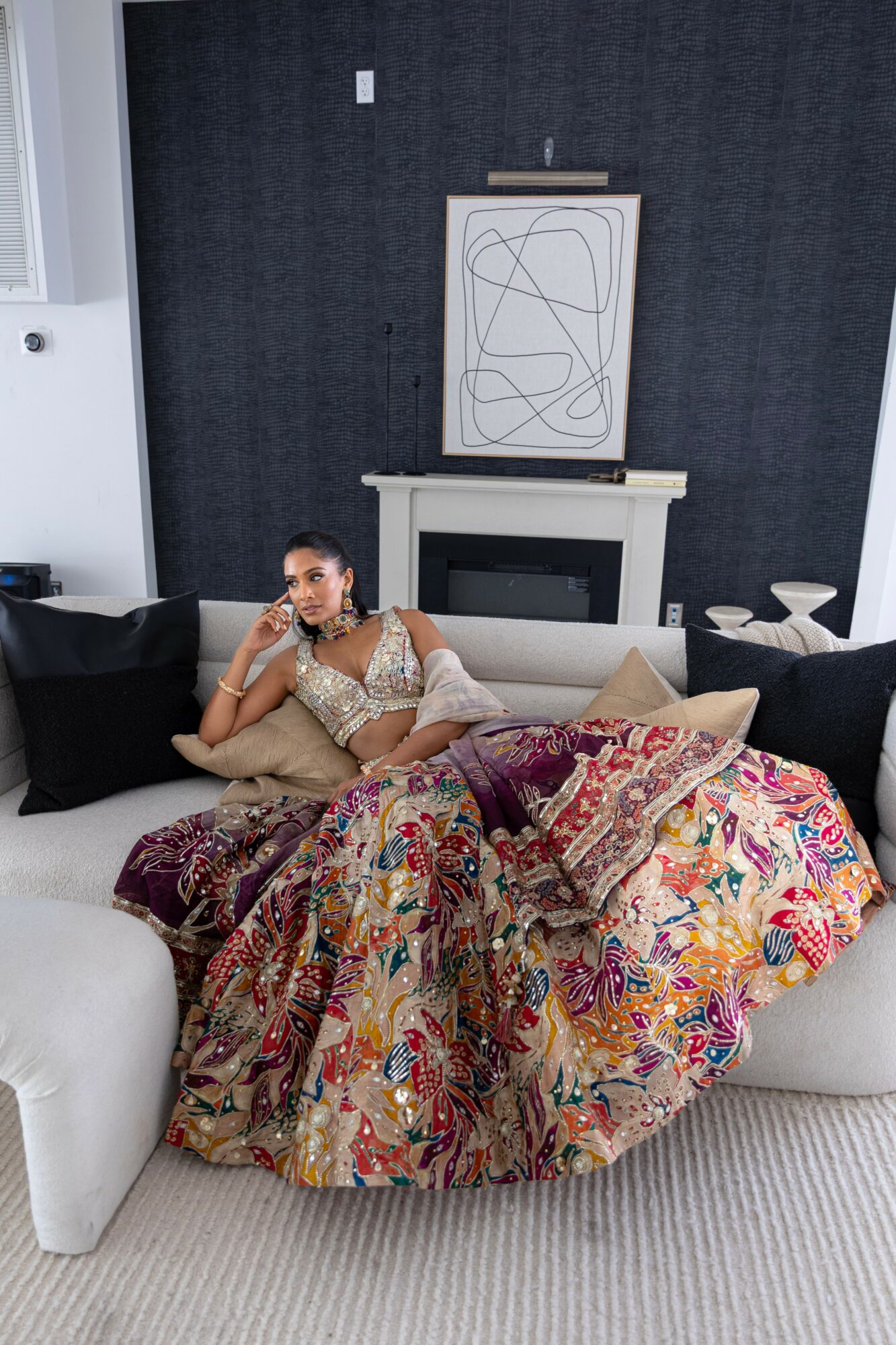Woman in colorful dress sitting on a sofa with black and beige cushions in a living room, with artwork and fireplace behind her.