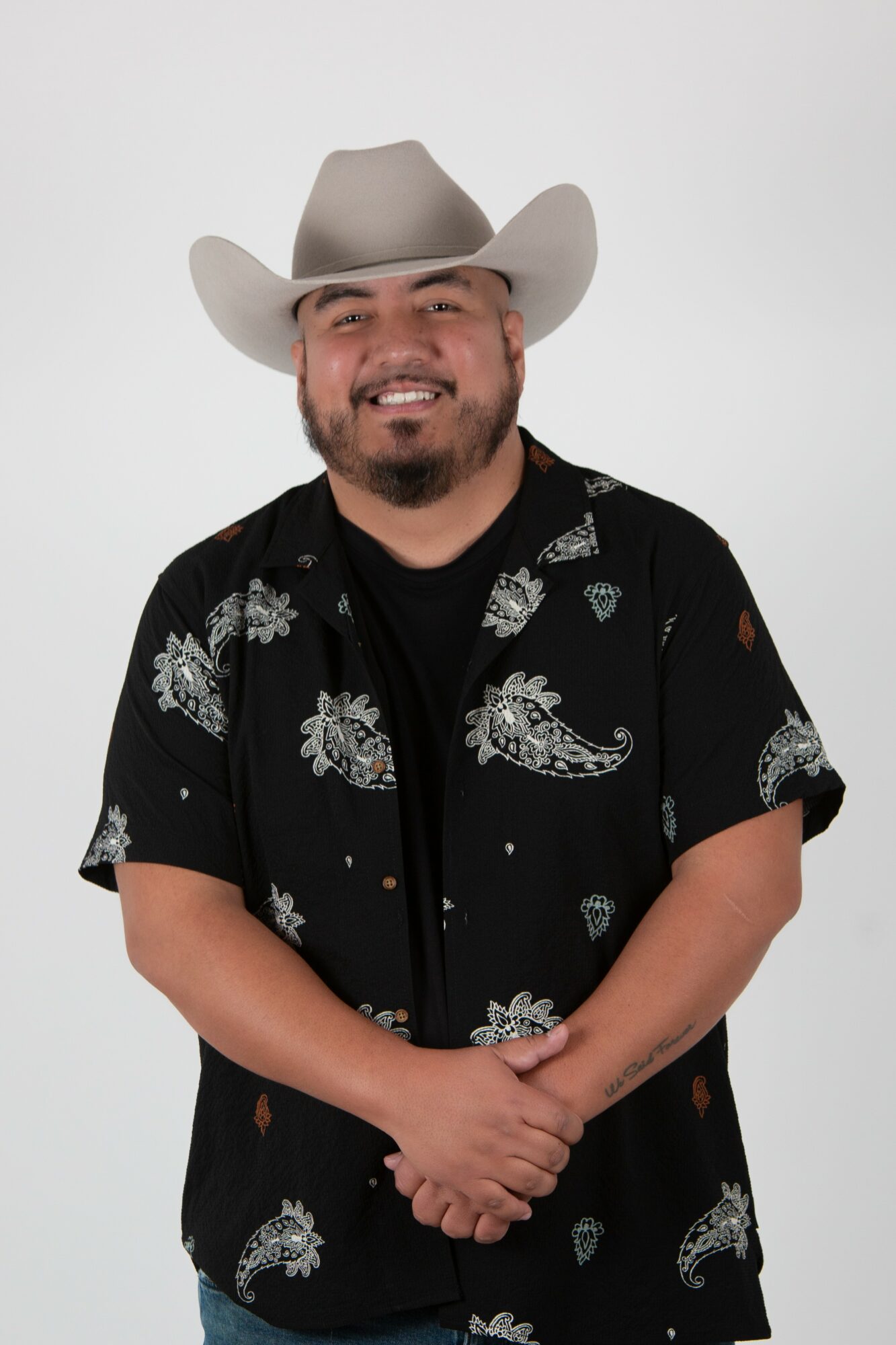Man with a beard and mustache wearing a cowboy hat and a black shirt with white floral patterns, smiling with arms crossed.