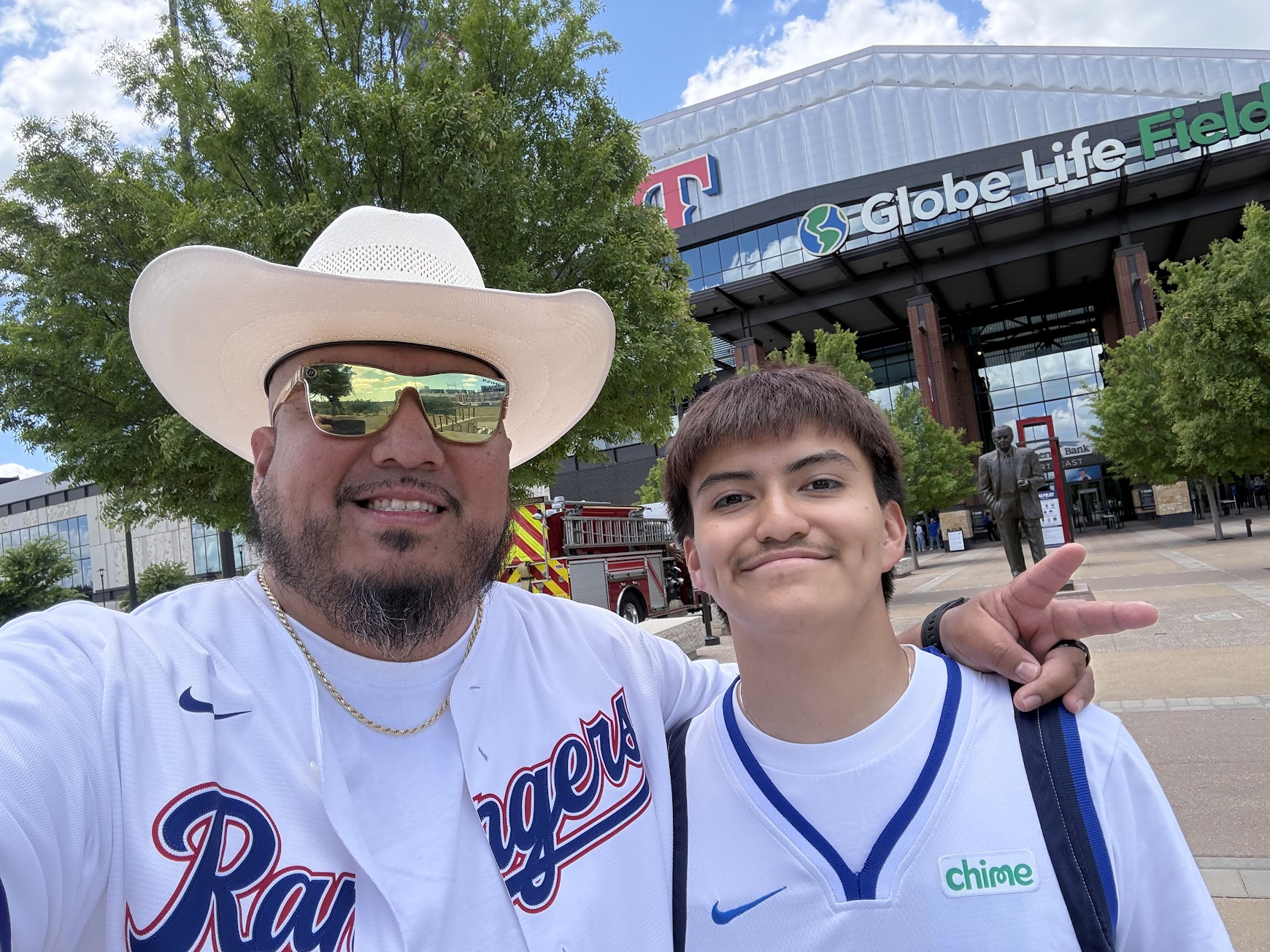 Two people smiling outdoors in front of a building with a sign, one wearing a cowboy hat and sunglasses, the other in a sports jersey.