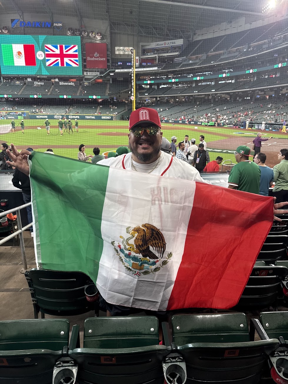 Person holding a Mexican flag at a baseball stadium, with a large screen displaying flags of Mexico and the UK in the background.