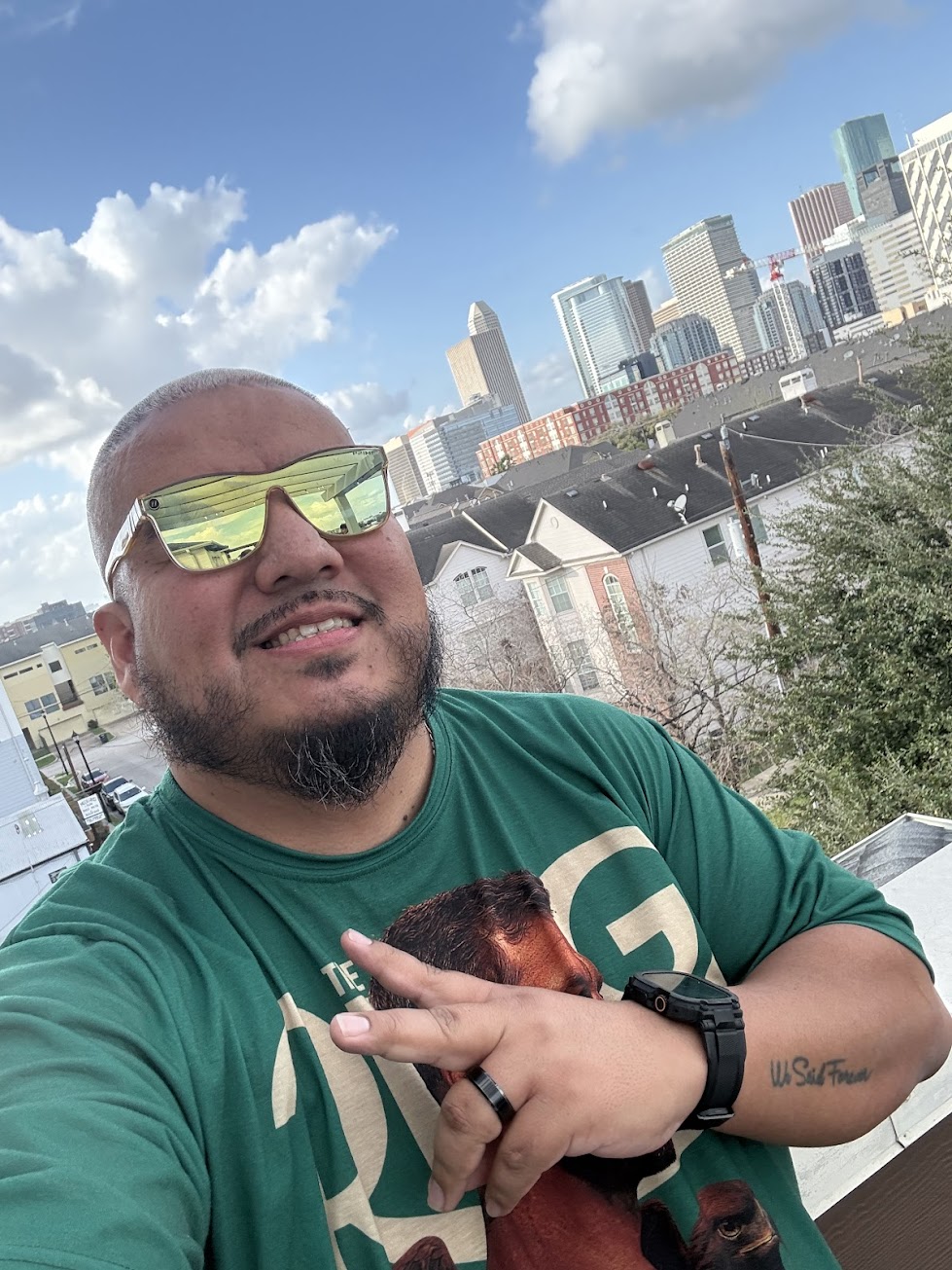 Man with sunglasses and a beard smiling outdoors with city skyline and blue sky in background.
