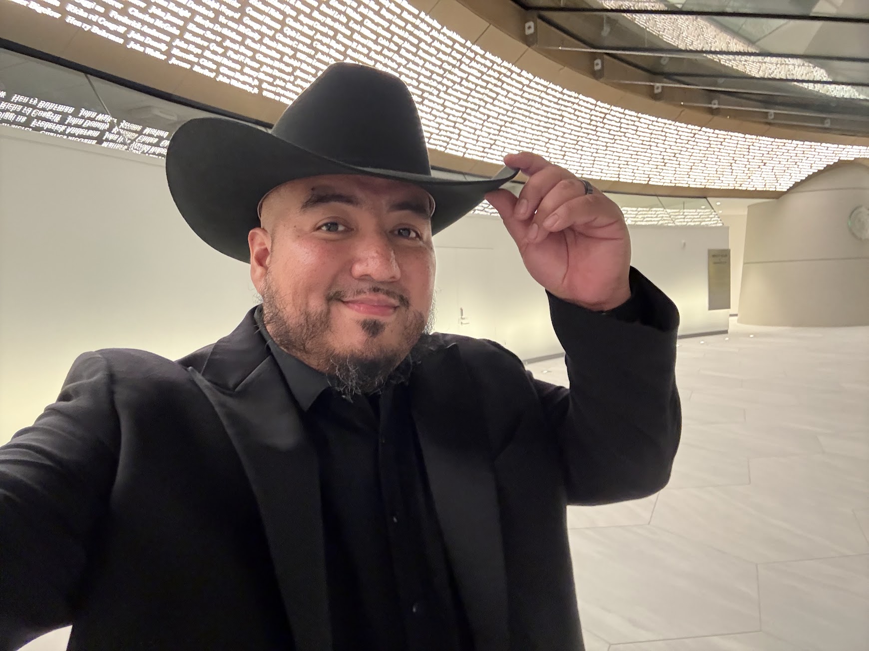 Man wearing a black hat and suit, smiling, in a modern indoor space with curved architecture.