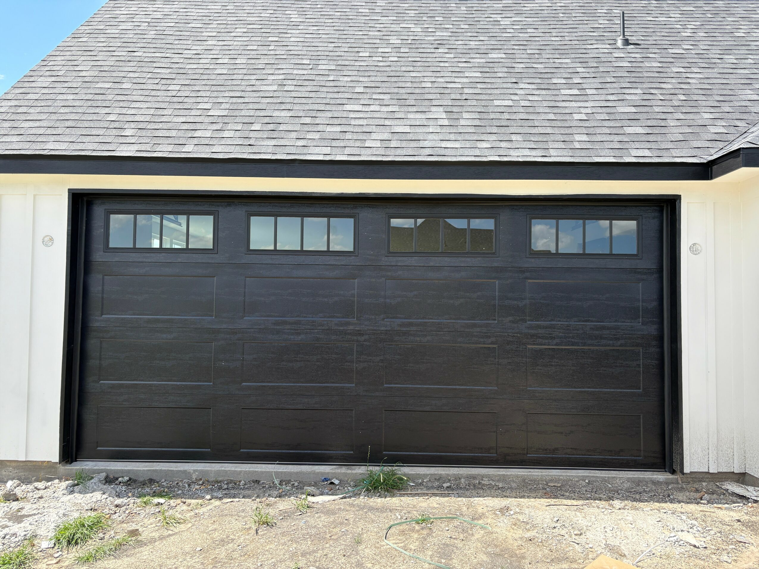 Black garage door with small rectangular windows at the top, part of a house with a gray shingled roof.