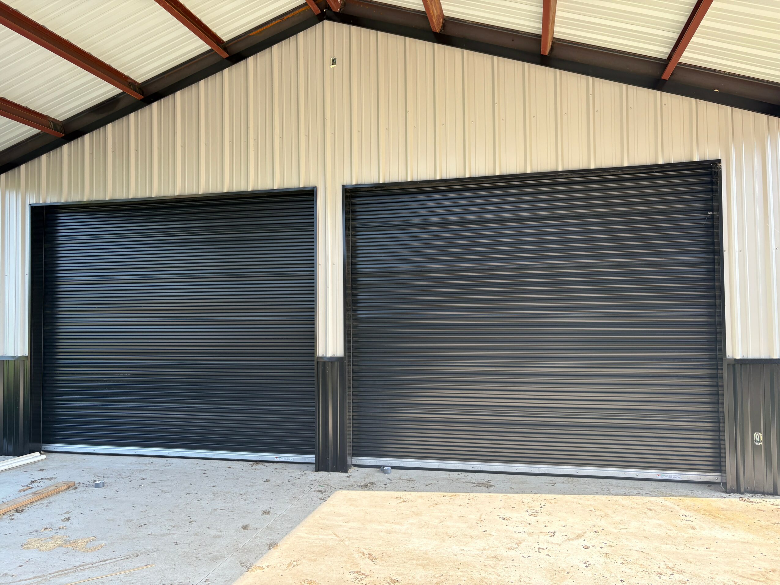 Two black garage doors on a beige building with a metal roof, concrete and dirt ground.
