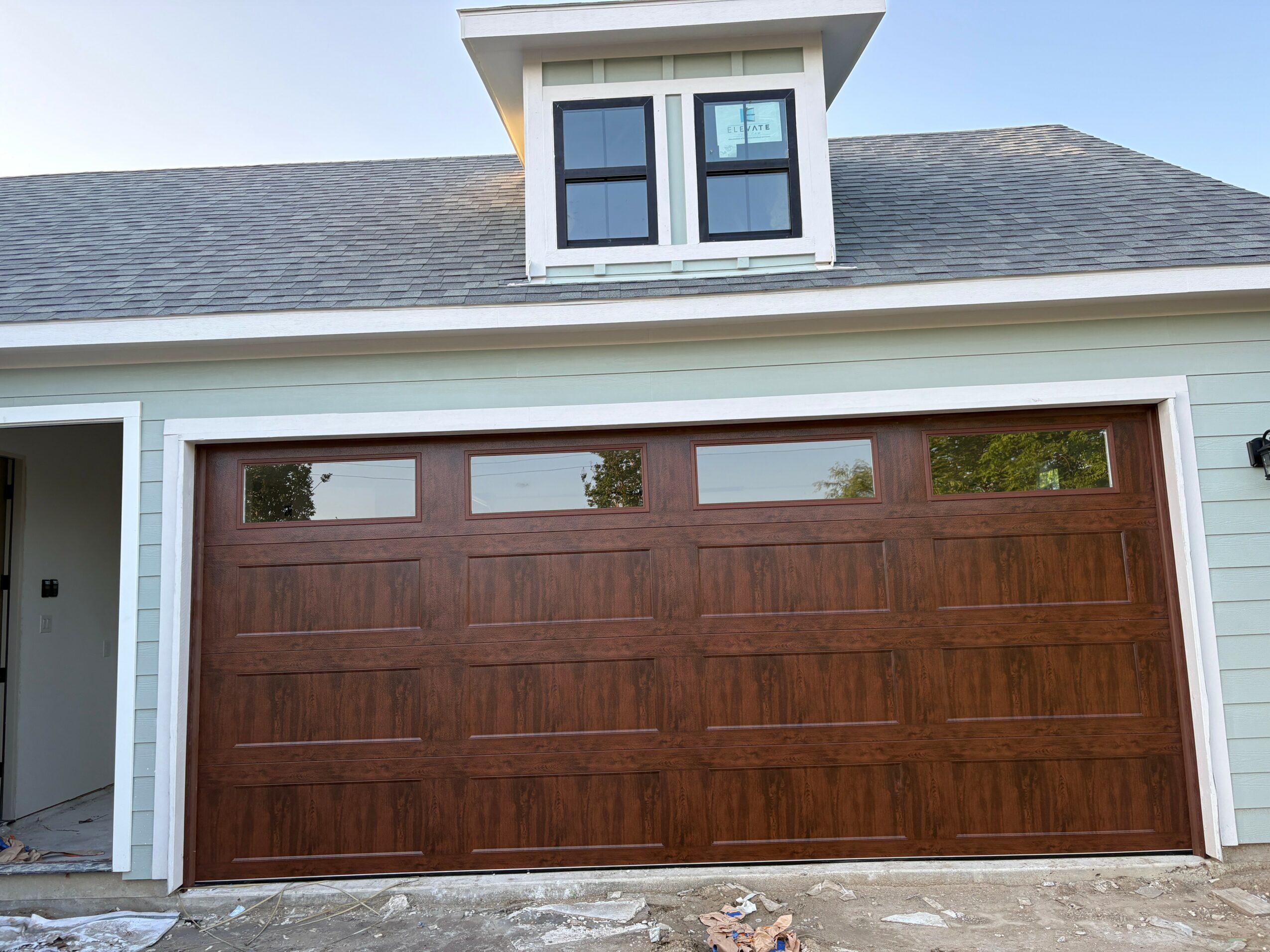 Wooden garage door with horizontal panels and small rectangular windows at the top, part of a house with white siding and a dormer window.