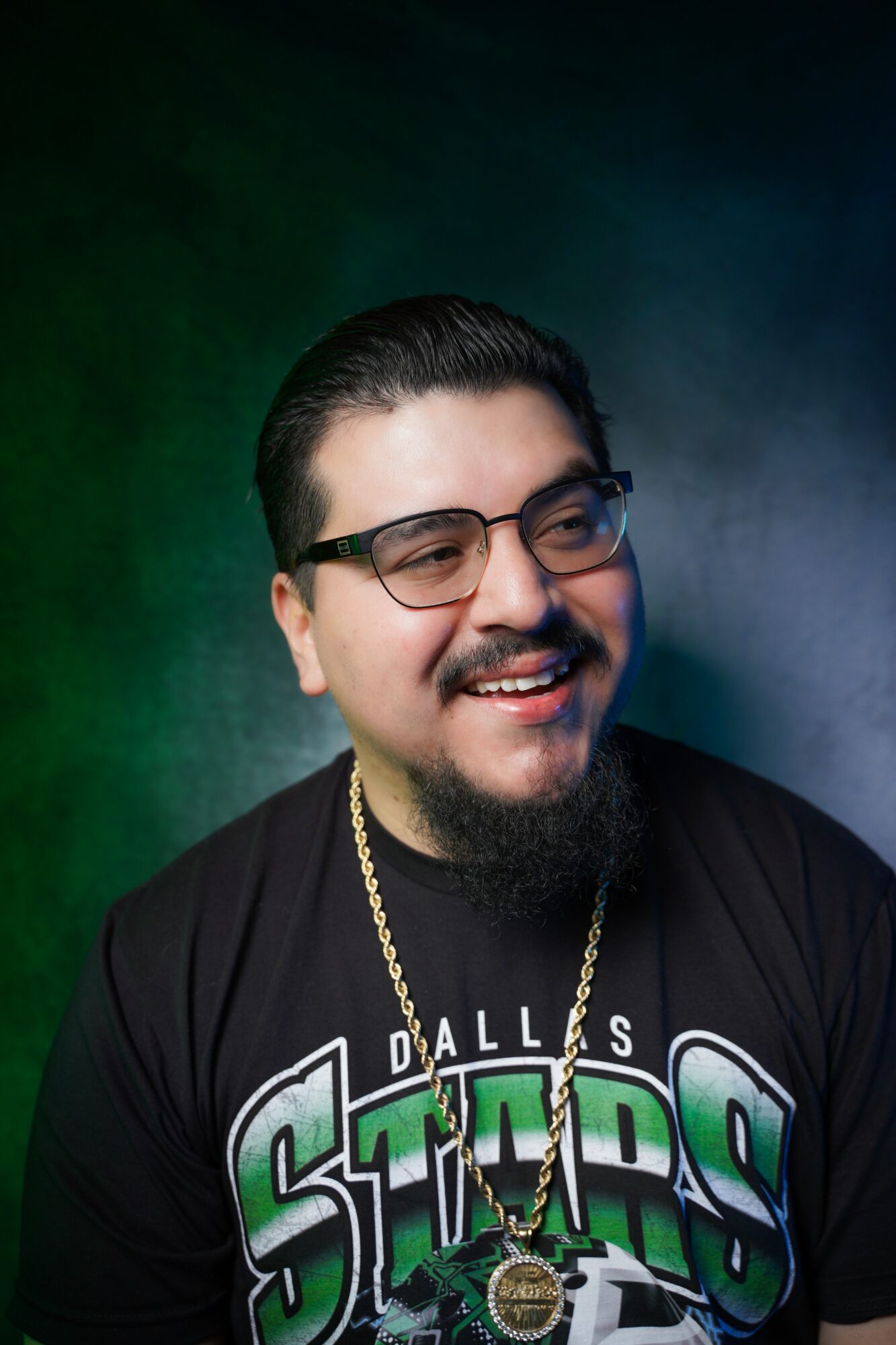 Smiling man with glasses, dark hair, beard, wearing a Dallas Stars shirt and gold chain, against a dark background.