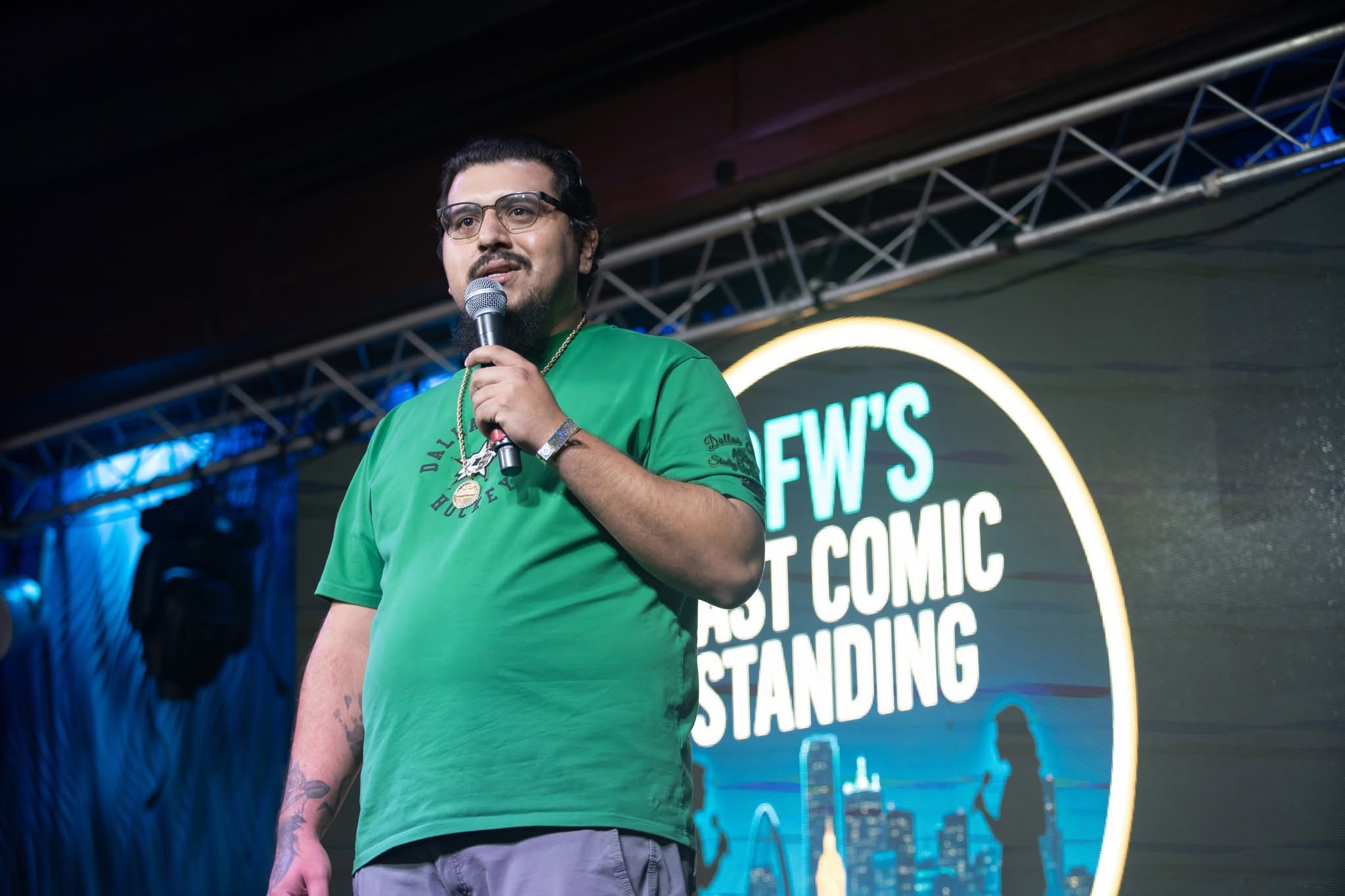 Person speaking into microphone on stage with a large circular sign behind them, wearing a green shirt.