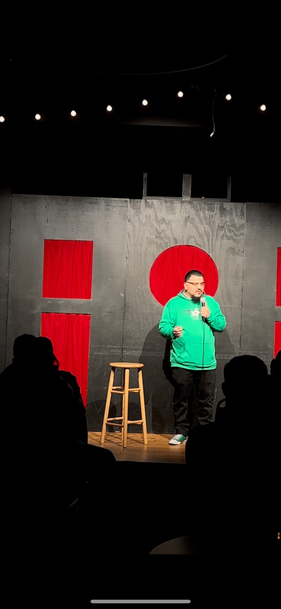 Person in green jacket on stage with red circle and square background, audience in foreground, stool nearby.