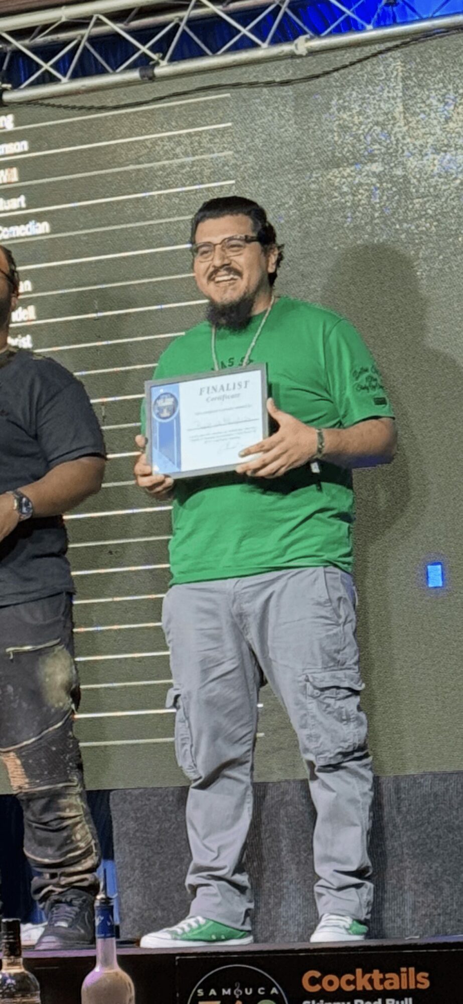 Person smiling holding a certificate on stage, wearing a green shirt and gray pants, with a large screen behind him.