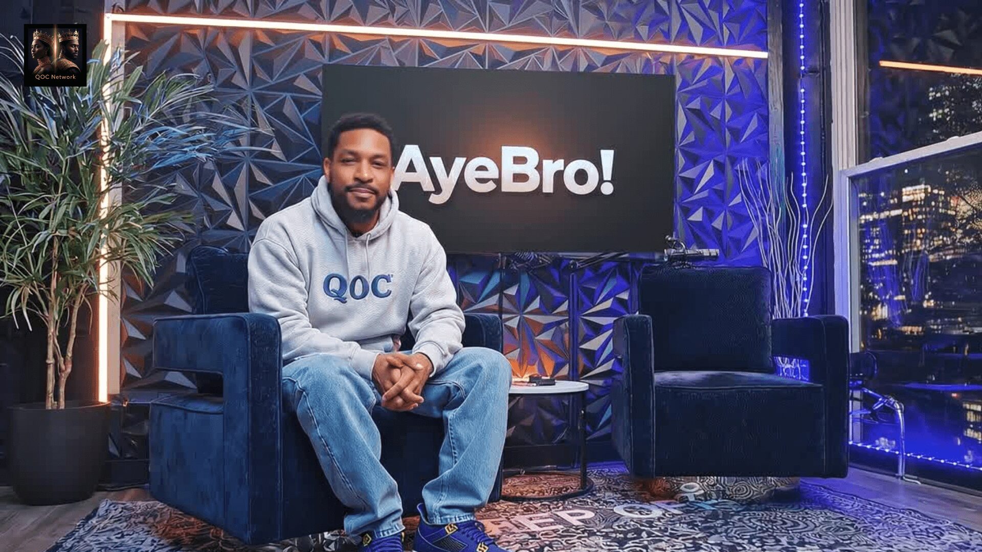 Man sitting on a blue sofa in a studio with a large screen behind him displaying 'AyeBro!'.