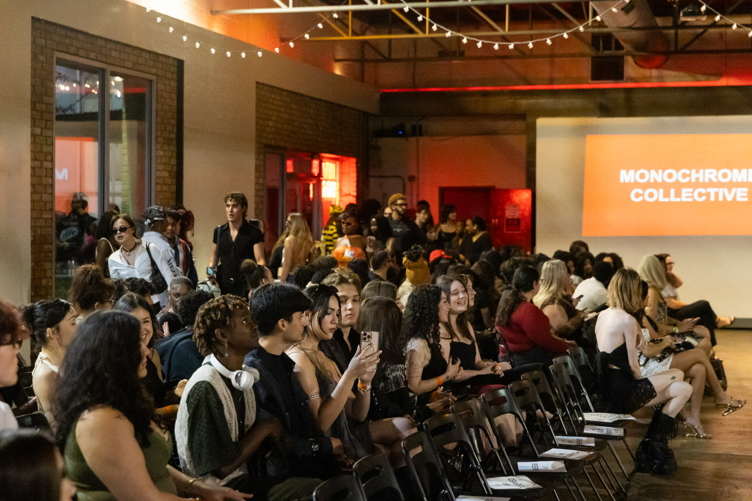 Audience seated in a large room watching a presentation on a screen, with some standing and socializing in the background.