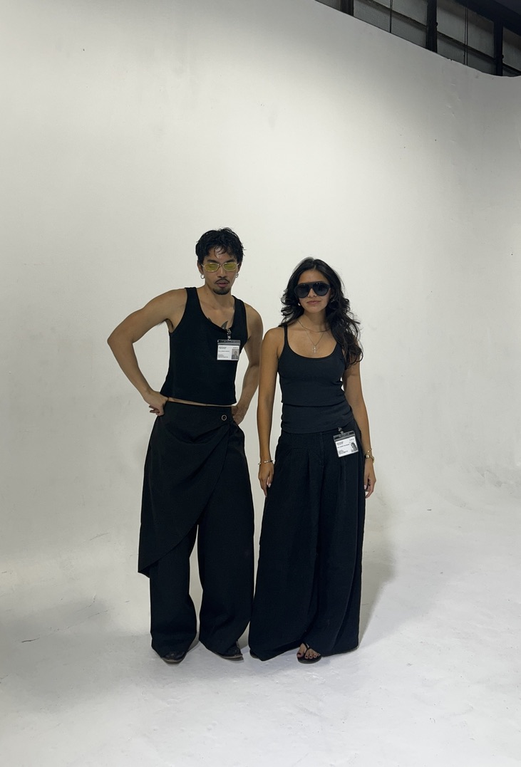 Two people stand against a plain white background, one wearing sunglasses and both dressed in dark clothing.