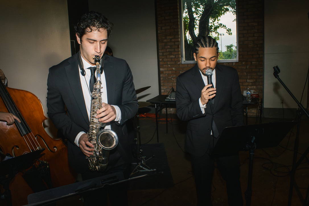 Two men in suits perform with microphones, one playing a saxophone, in an indoor setting with a window and brick wall.
