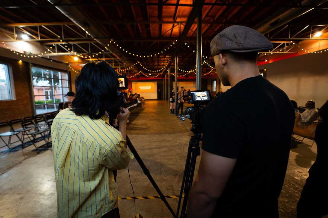 Two people filming in a large indoor space with string lights and seating, one with a camera and the other with a microphone.