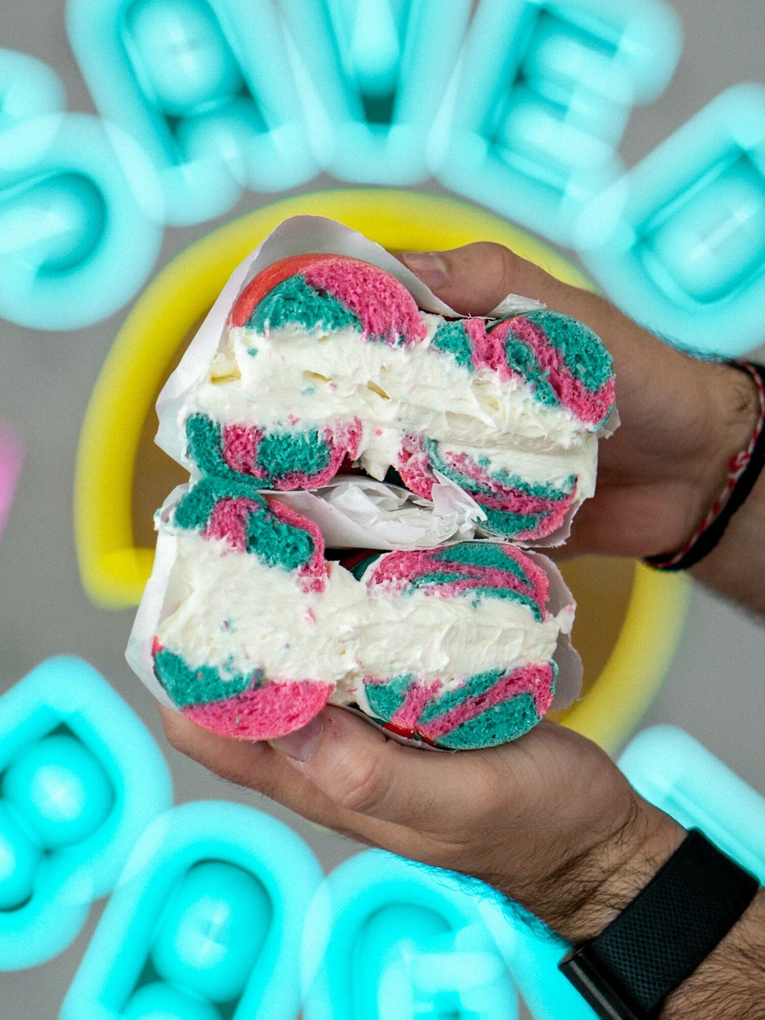 Person holding a colorful ice cream sandwich with pink, blue, and white swirls, neon signs in the background.