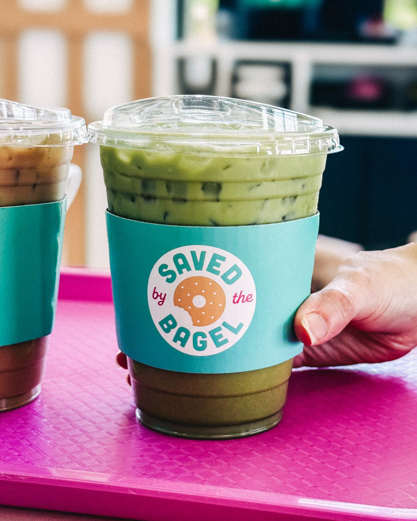 Clear plastic cup with green and brown layered drink, labeled 'Saved by the Bagel,' held by a person's hand on a pink surface.