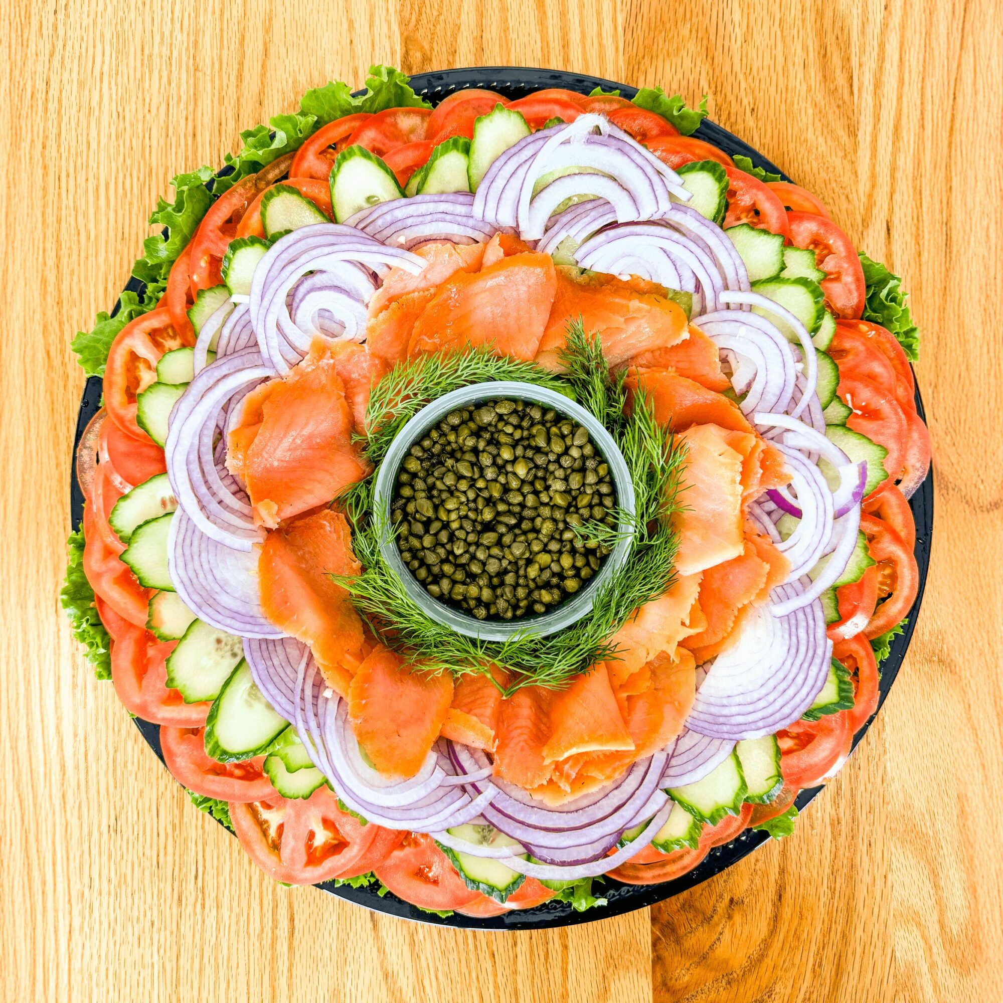 A round salad with sliced tomatoes, cucumbers, onions, and smoked salmon, garnished with herbs and a small bowl of green sauce in the center.