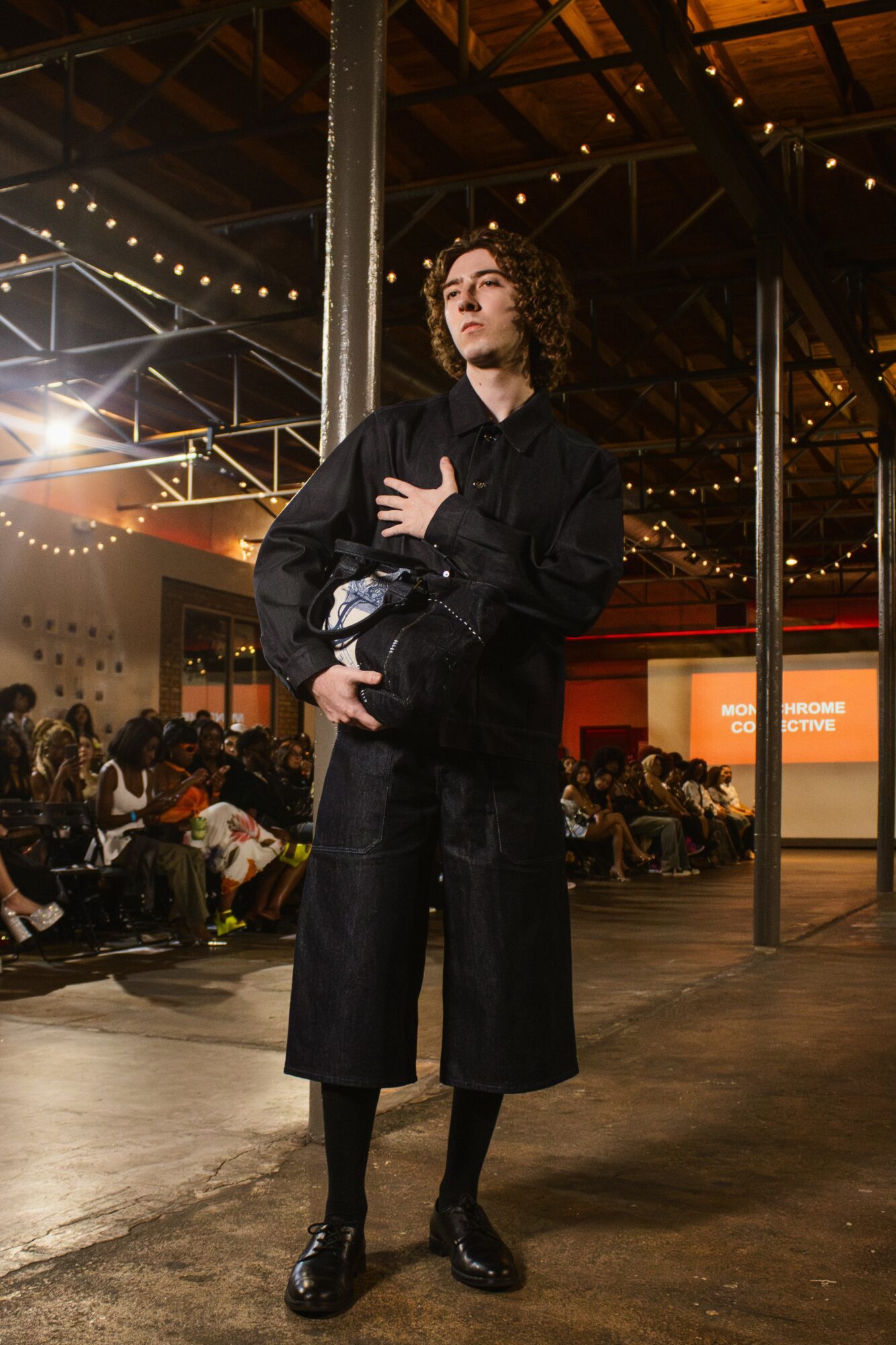 Person with curly hair standing in a large indoor space with string lights and seated audience, wearing dark clothing and holding a bag.