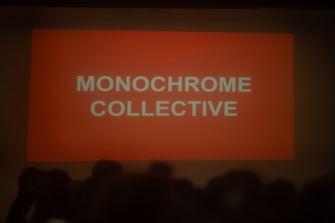 Red screen with white text reading 'MONOCHROME COLLECTIVE' in a dark room with silhouettes of audience members.