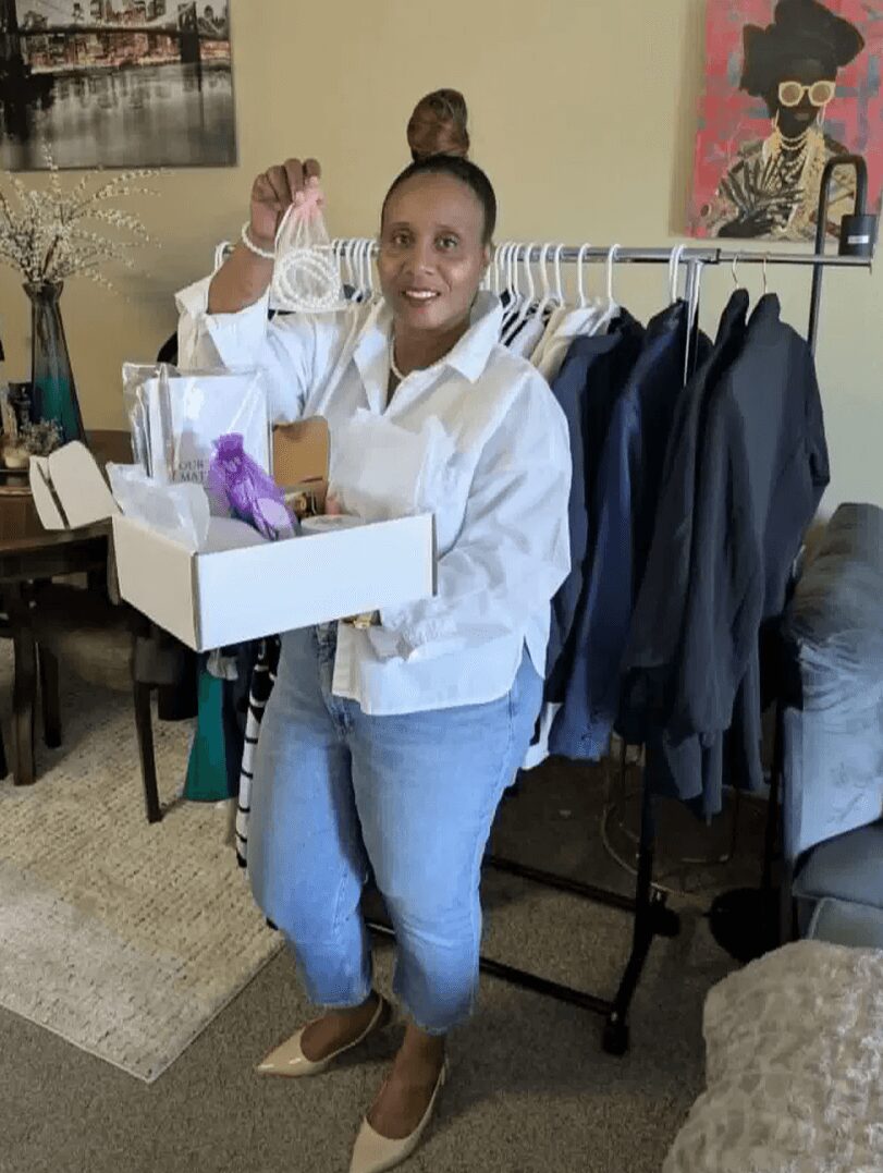 Person holding a small item in a room with clothing rack and artwork, smiling, wearing a white shirt and jeans.