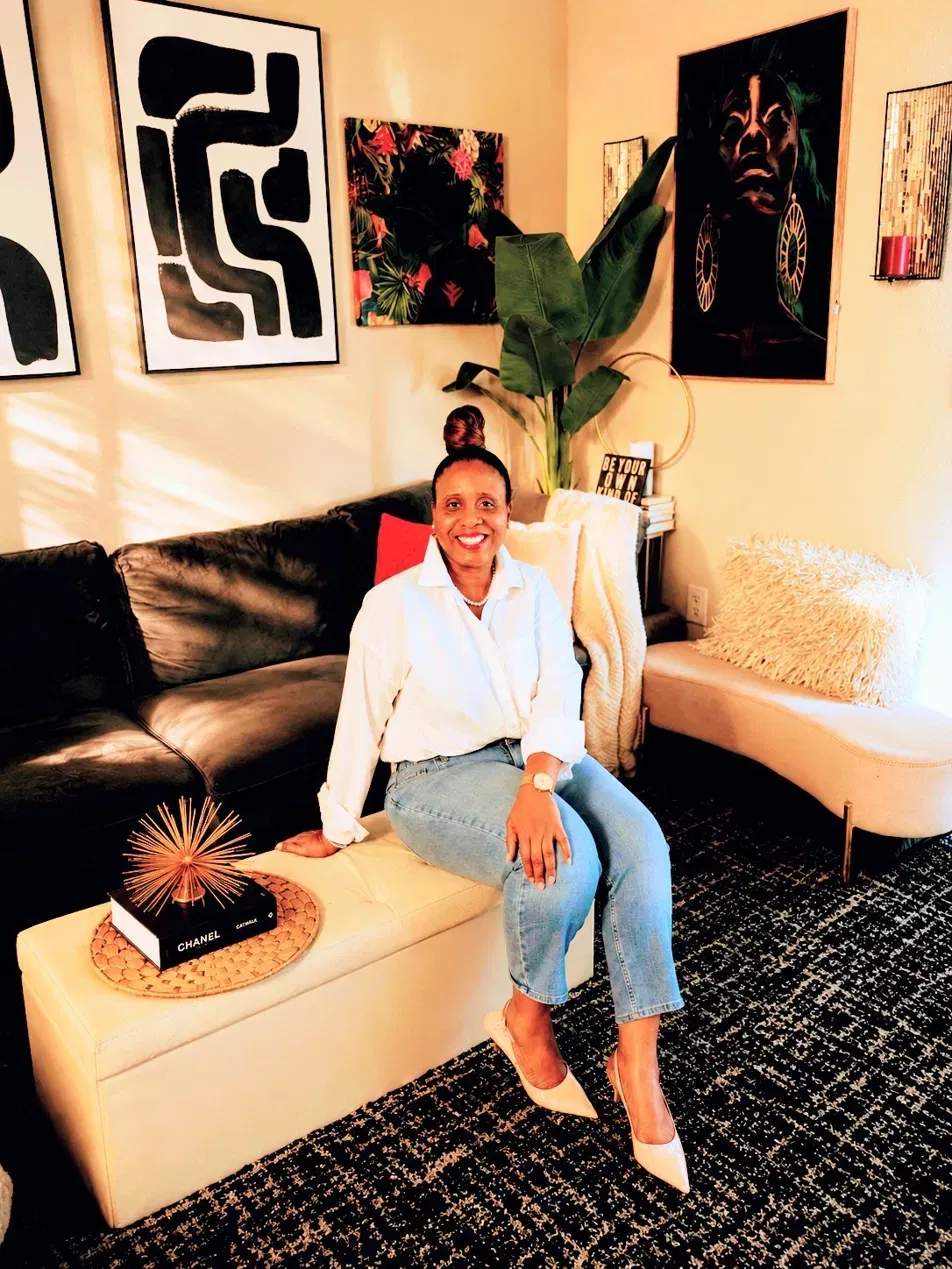 Woman sitting on a beige sofa in a room with artwork and plants, smiling, wearing a white shirt and jeans.
