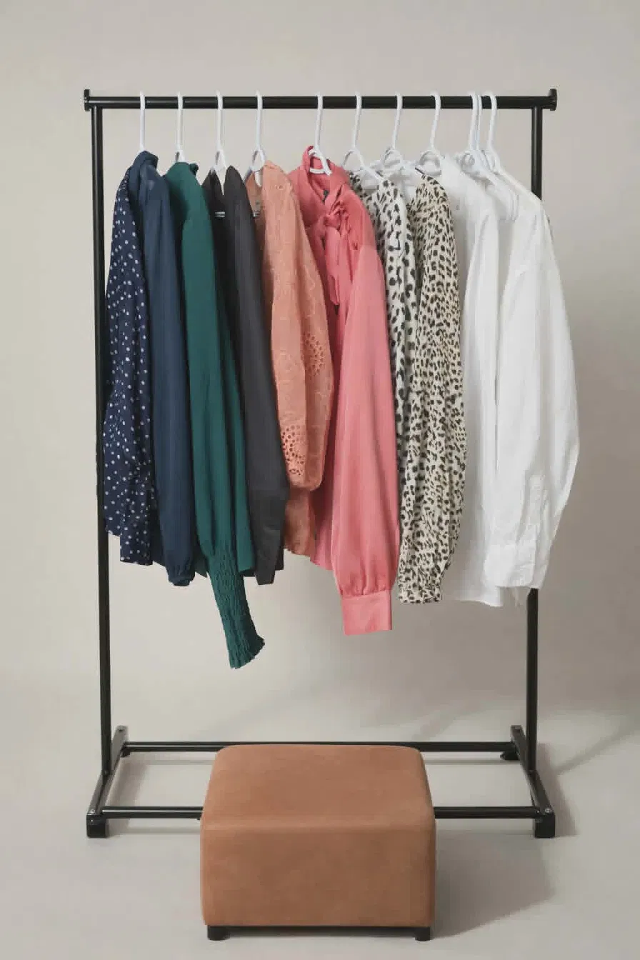 Clothing rack with various jackets and shirts, and a brown ottoman at the bottom, against a plain background.