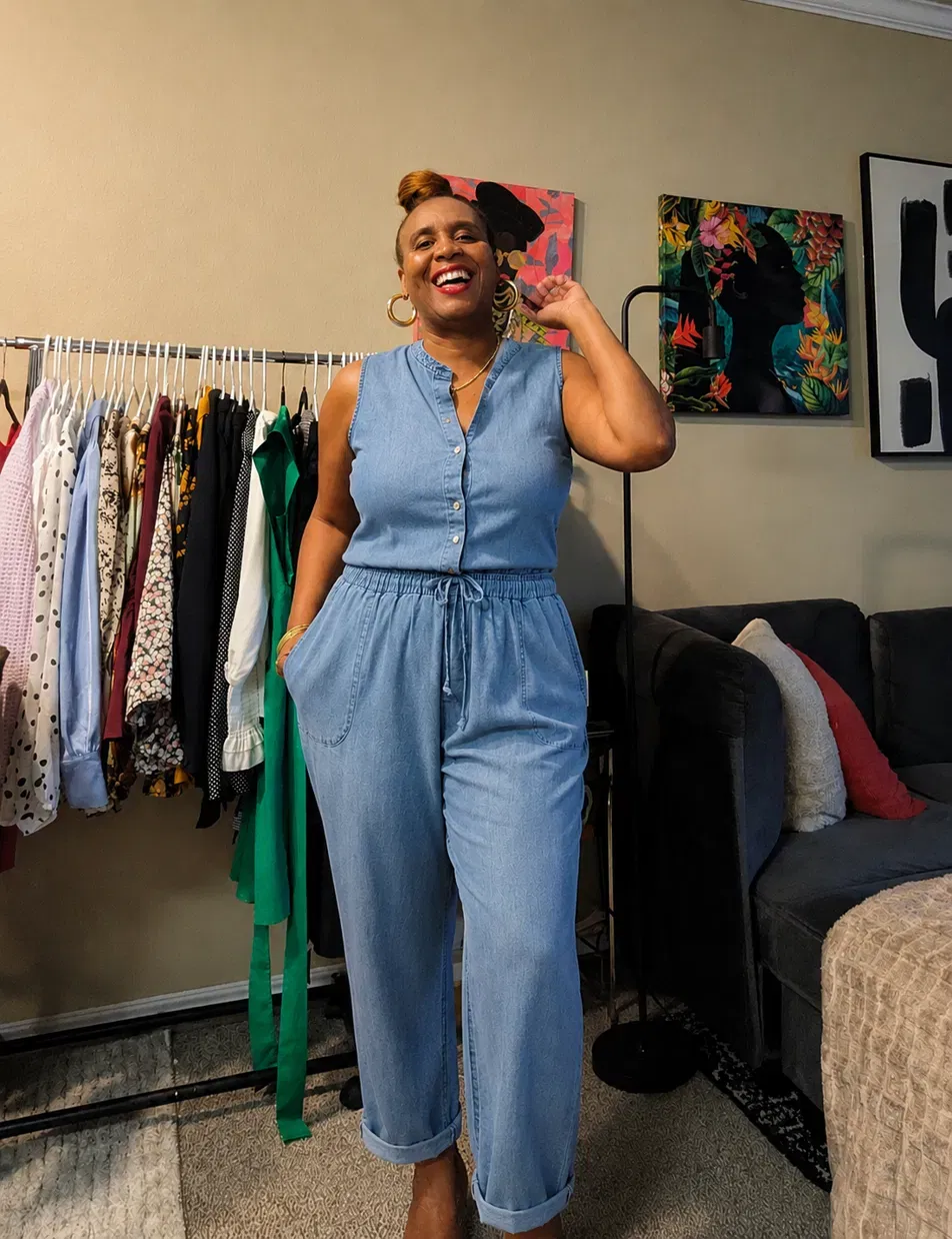 Smiling woman in denim jumpsuit standing in a clothing store with colorful artwork on the wall behind her.