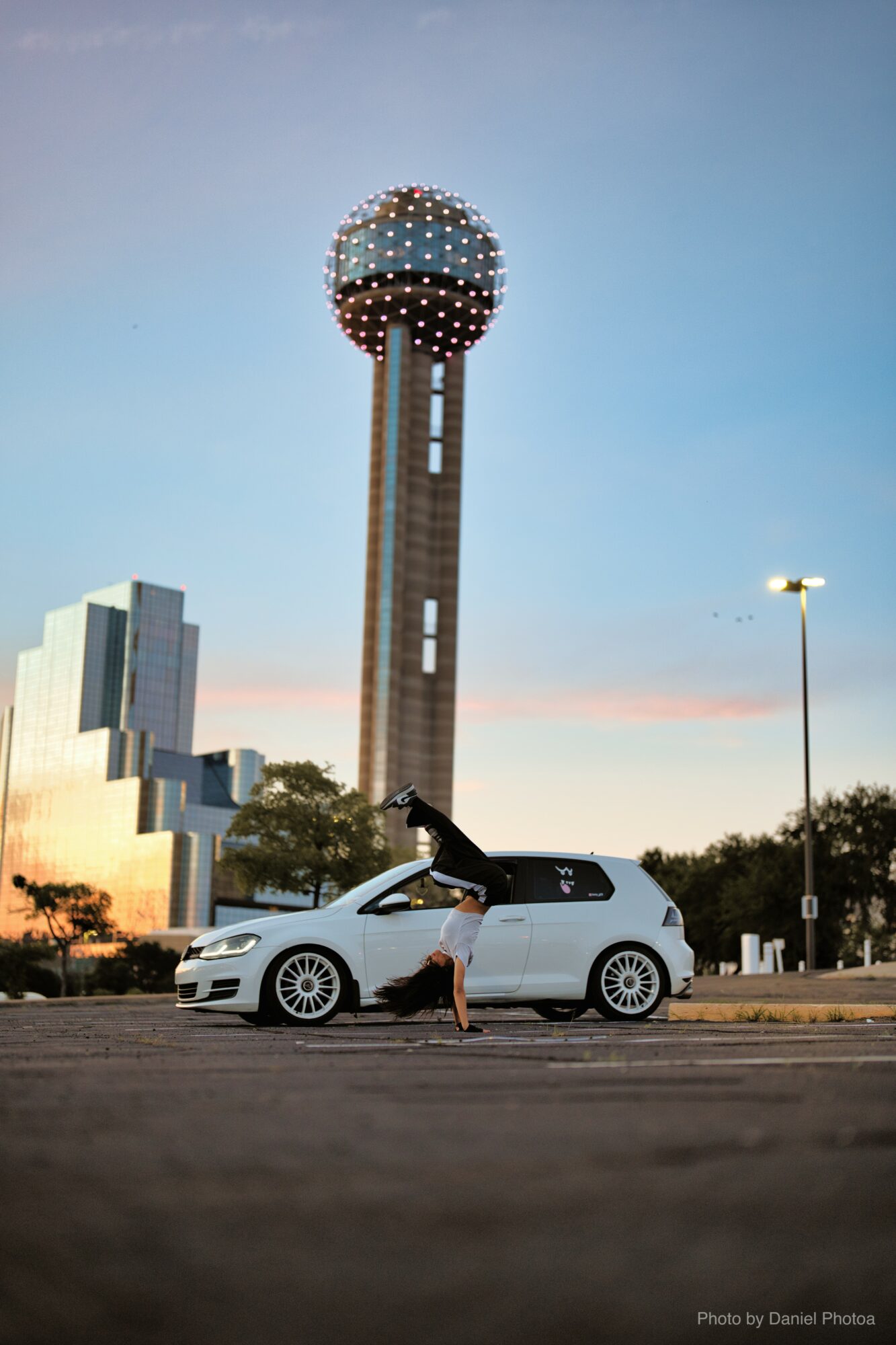 Person standing next to a white car in front of a tall tower with a spherical top, against a sky with sunset colors.