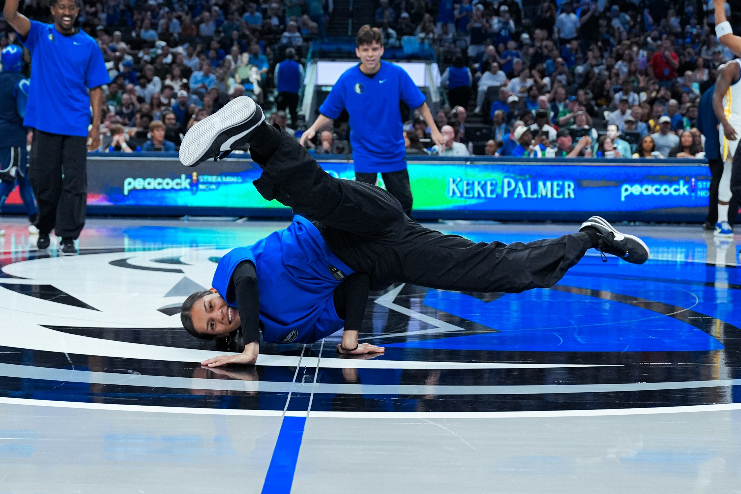 Person in blue shirt performing a breakdancing move on a basketball court, with spectators in the background.