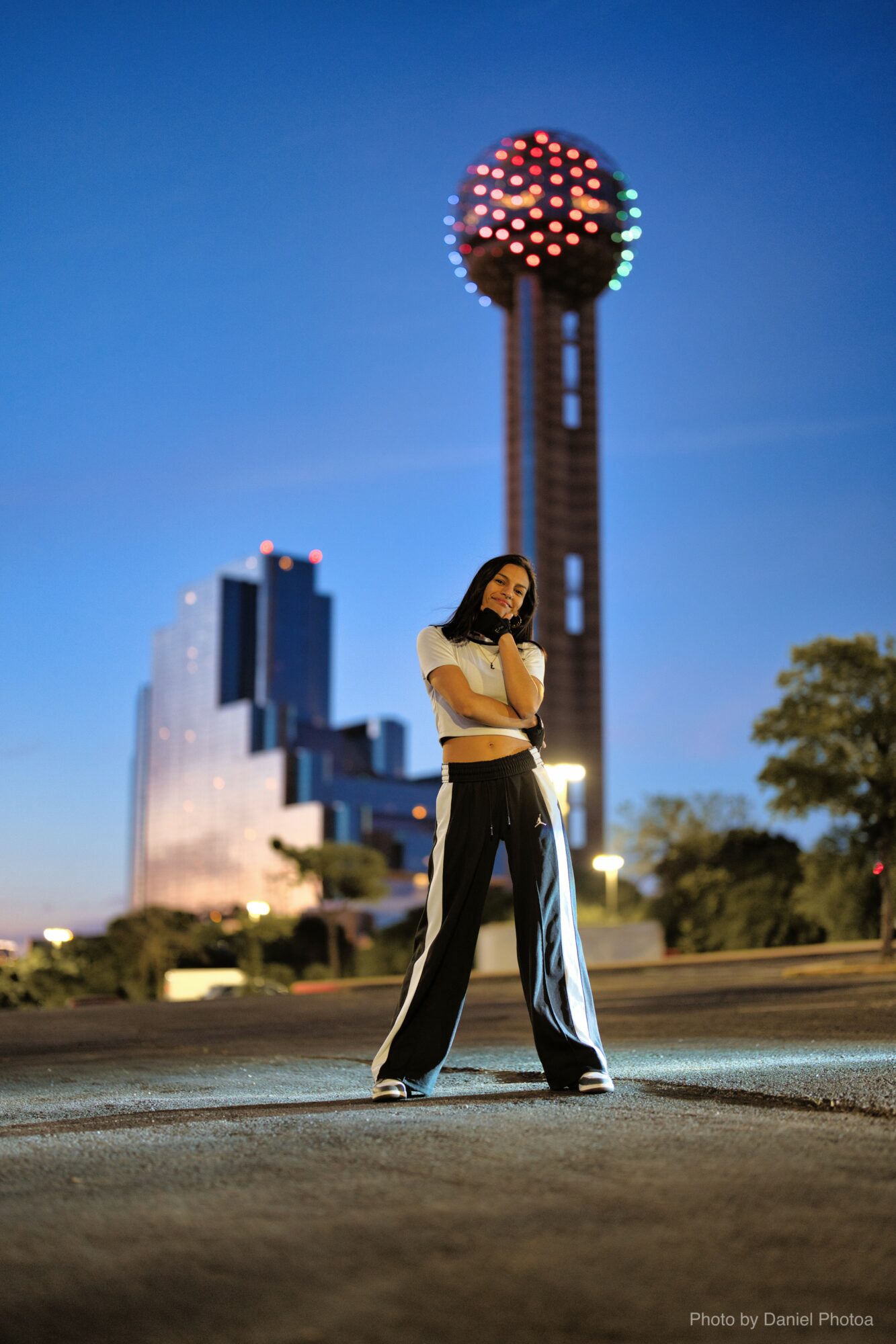 Young woman standing outdoors at night with a cityscape and illuminated tower in background, arms crossed, looking at camera.