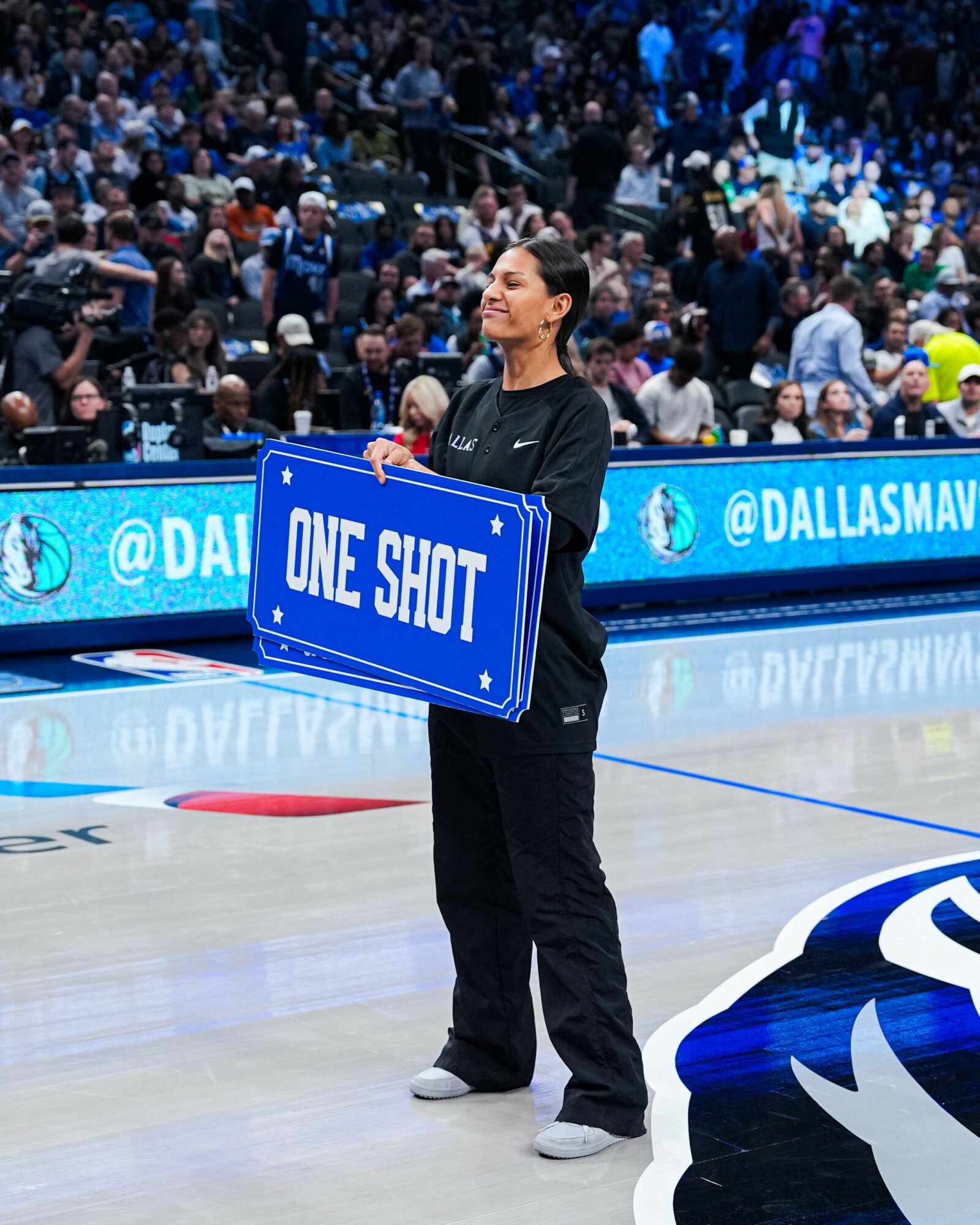 A woman holding a blue sign with white text on a basketball court, with a crowd in the background.