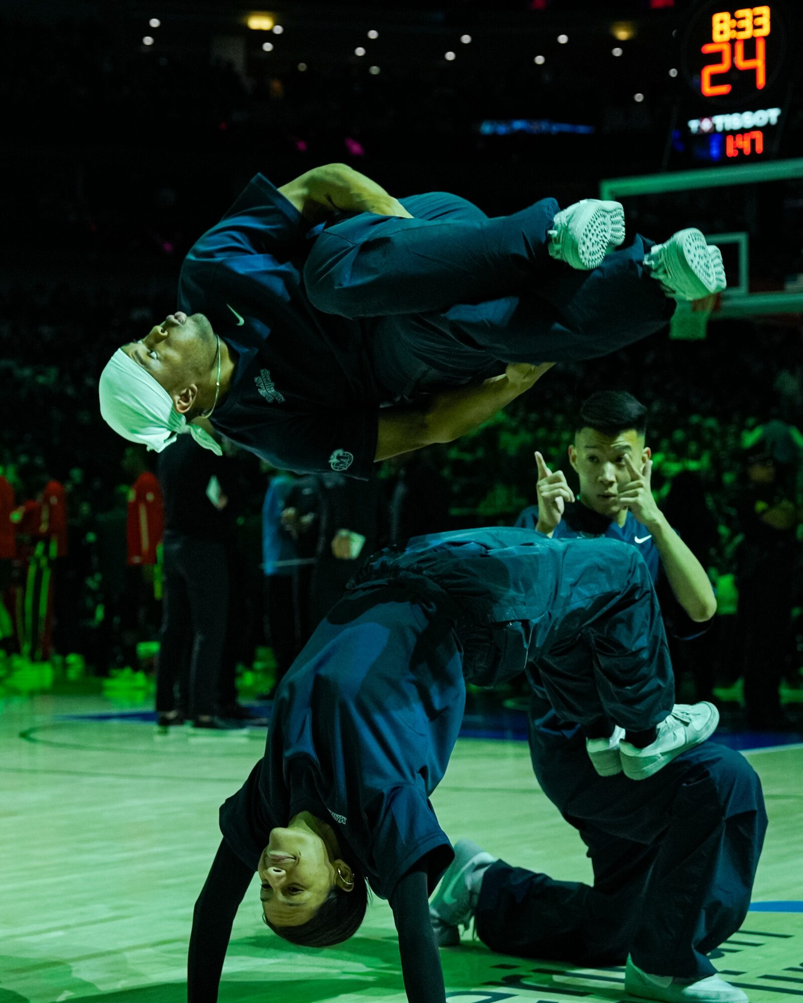 Two performers in dark clothing and white head coverings perform a balancing act on a basketball court, with a scoreboard in the background.