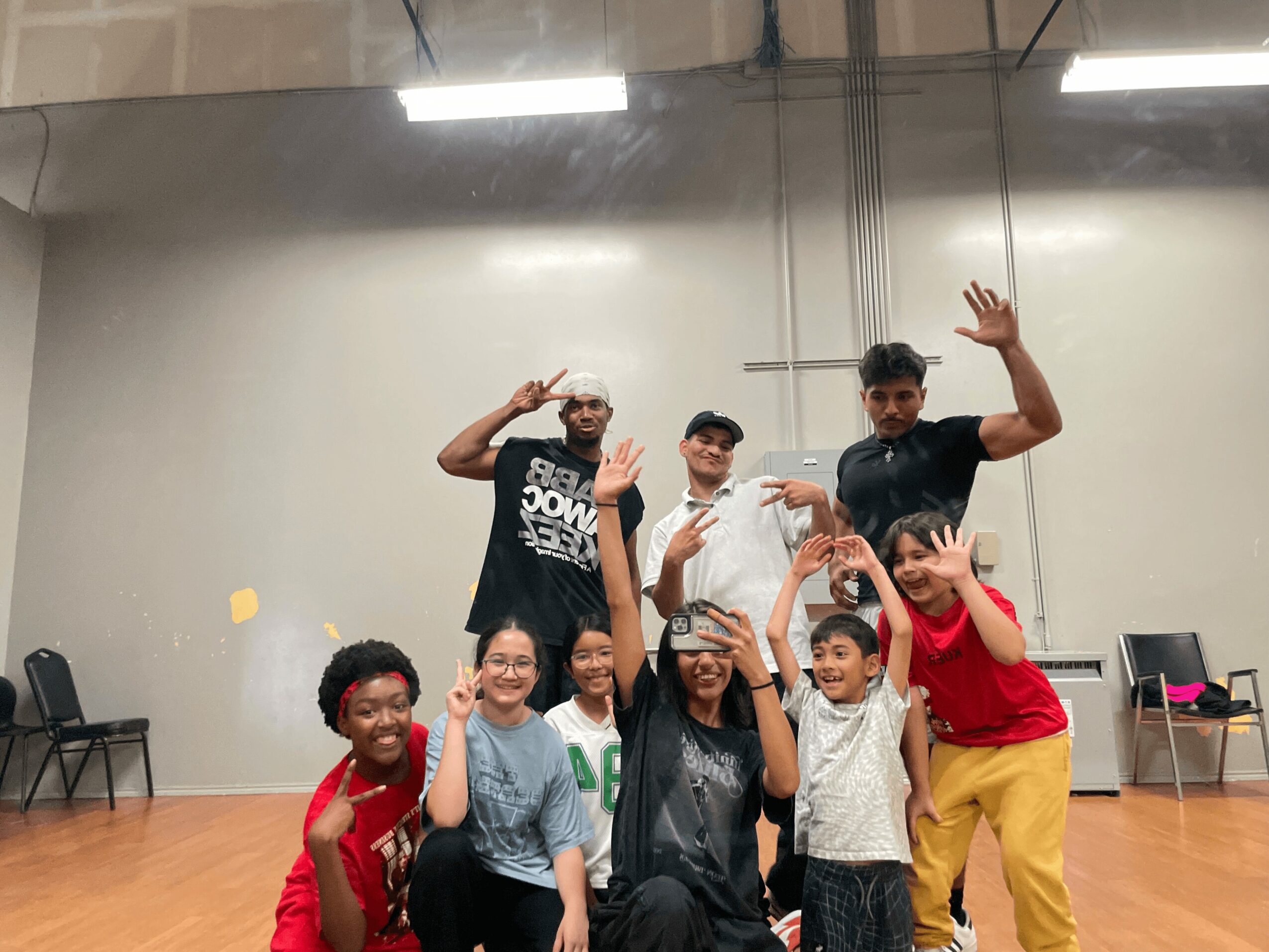 Group of seven young people posing indoors, some making peace signs and waving, in a spacious room with chairs and a gray wall.