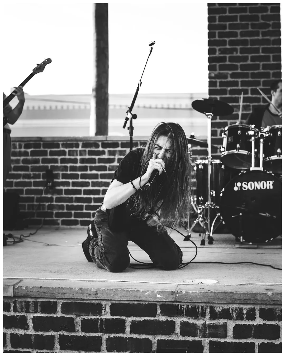 Kneeling woman singing into a microphone on stage with a drum set and brick wall background.