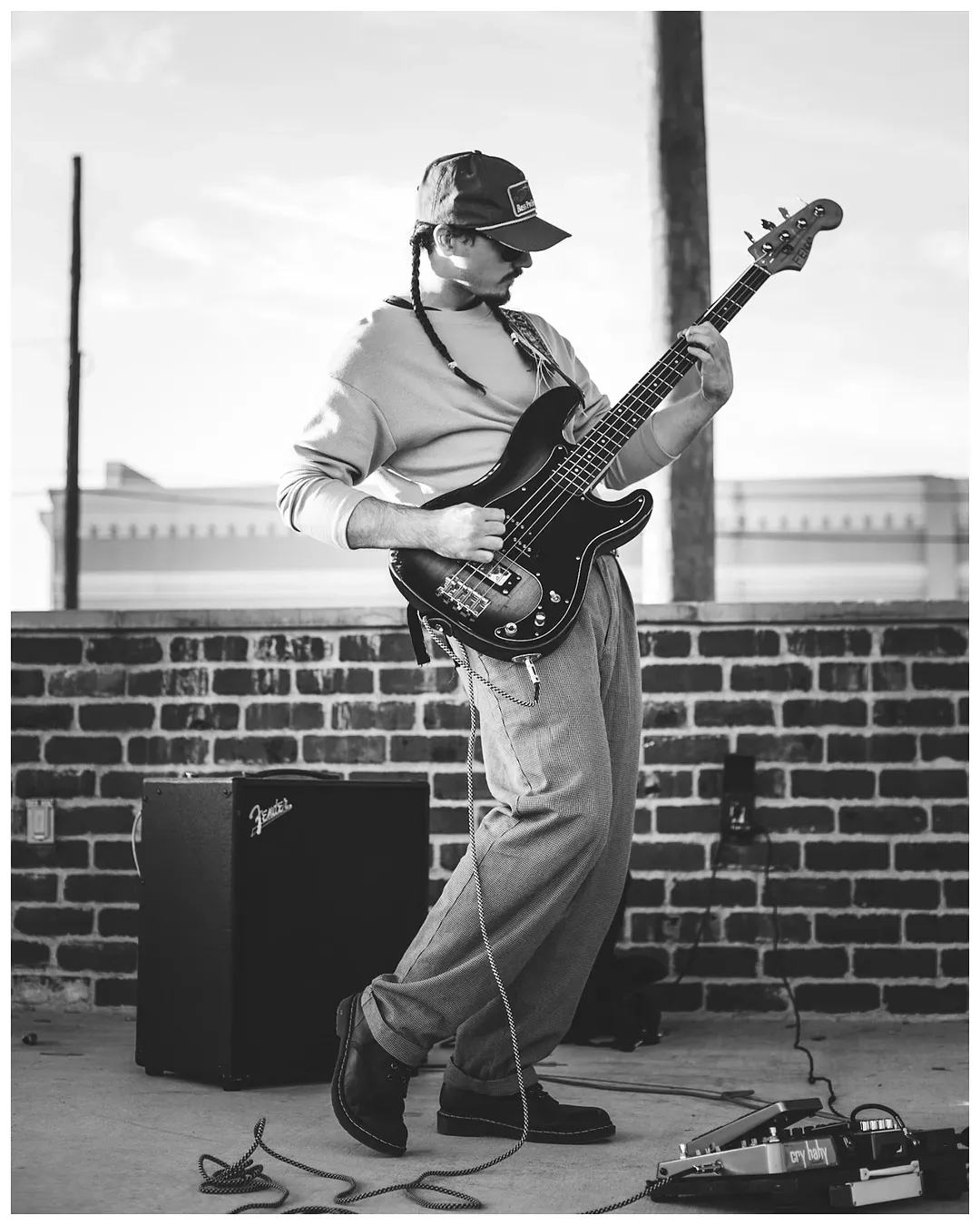 Person playing electric guitar outdoors on a brick wall background, wearing a cap and casual clothing.
