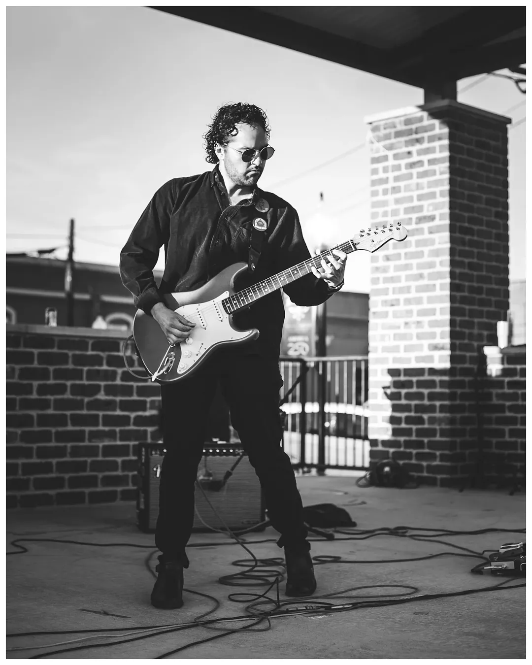 Man playing electric guitar outdoors on a rooftop with brick structures, wearing sunglasses and a dark jacket.