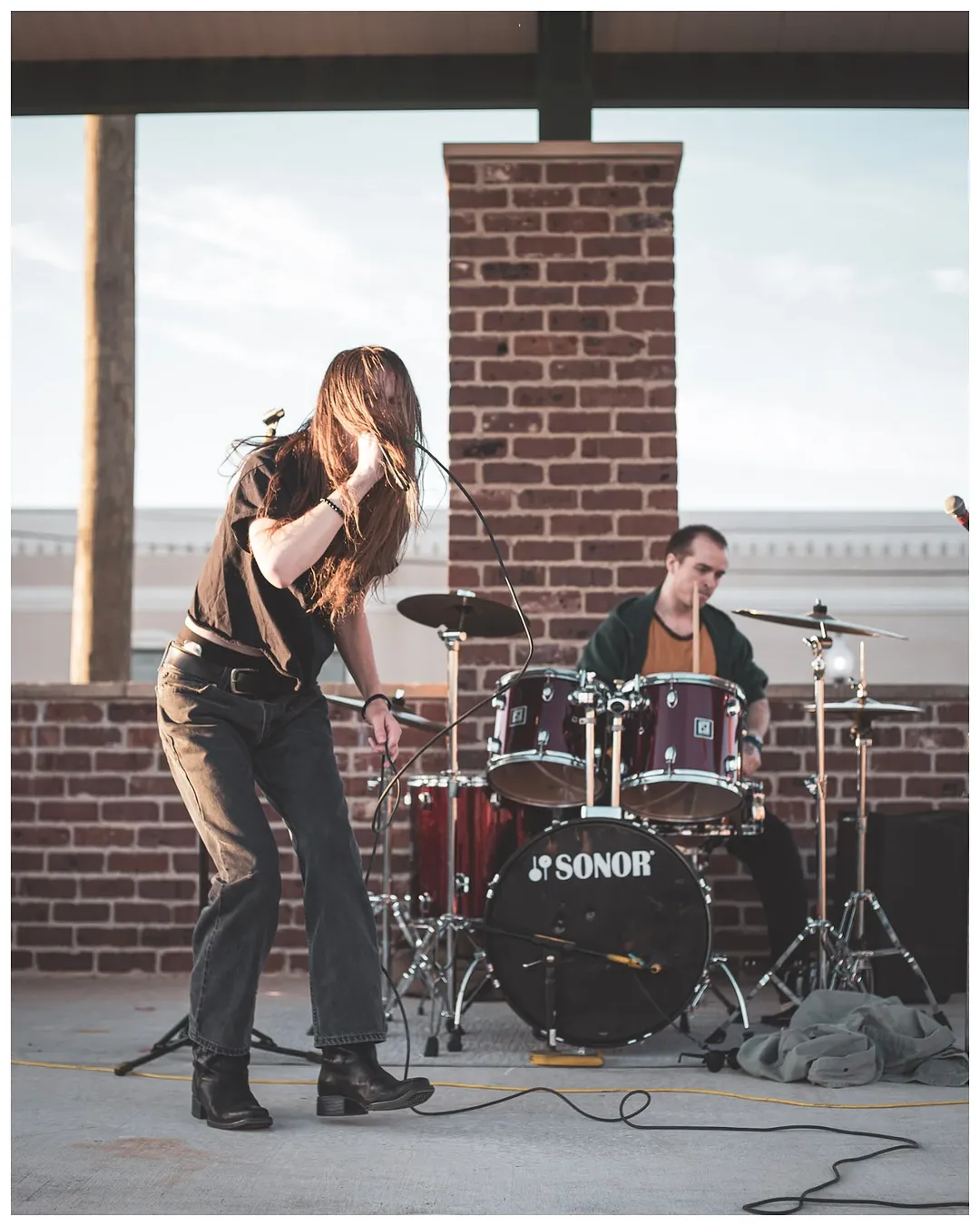 Two musicians perform outdoors under a bridge, one singing and the other playing drums, with a brick wall background.