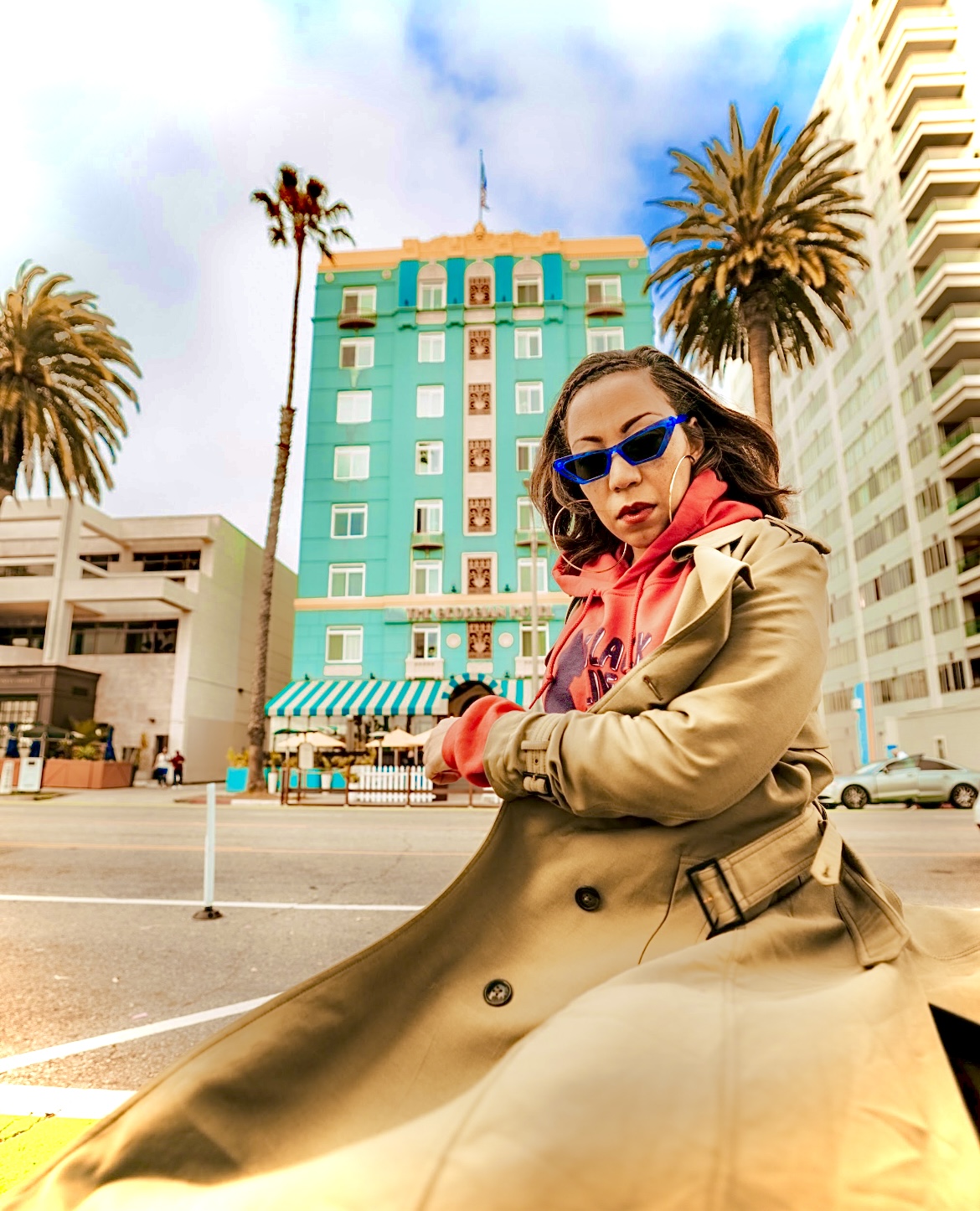 Woman wearing sunglasses and a beige trench coat sitting outdoors with tall palm trees and a blue building in the background.