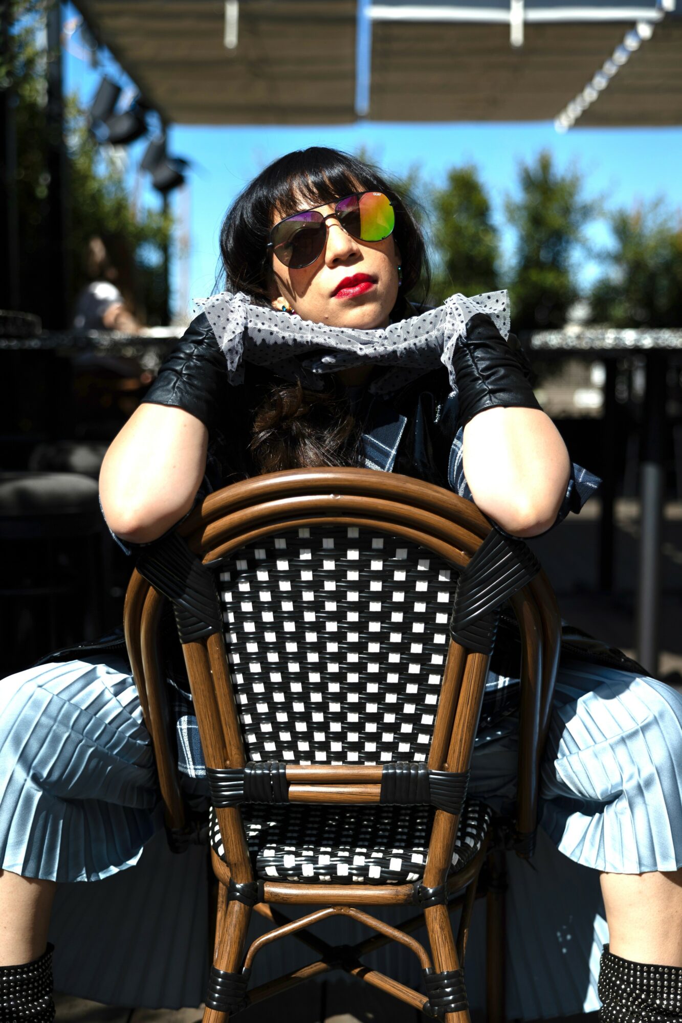 Woman with sunglasses and black gloves sitting outdoors, holding a scarf, with trees and sky in background.